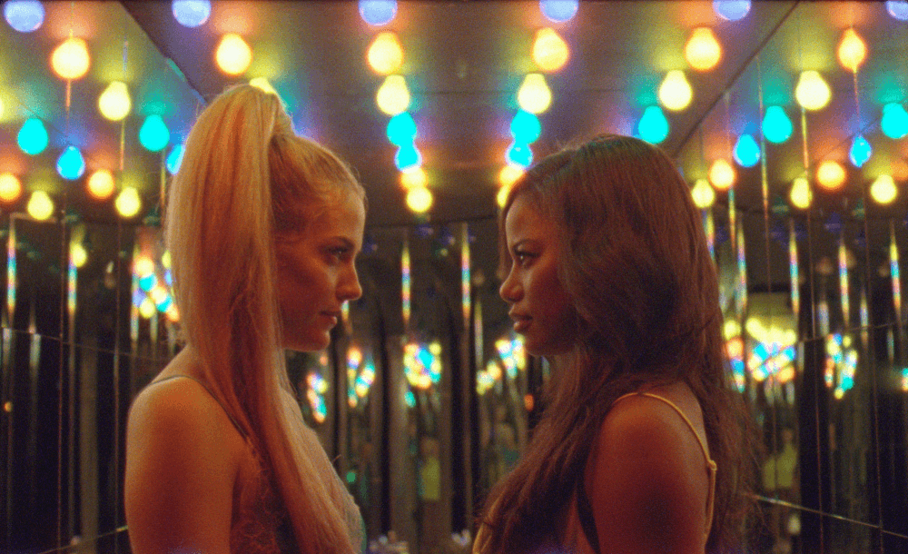 Still from the film "Zola" shows the side profiles of two women as they face each other. Behind them, rows of rainbow light bulbs line the ceiling.