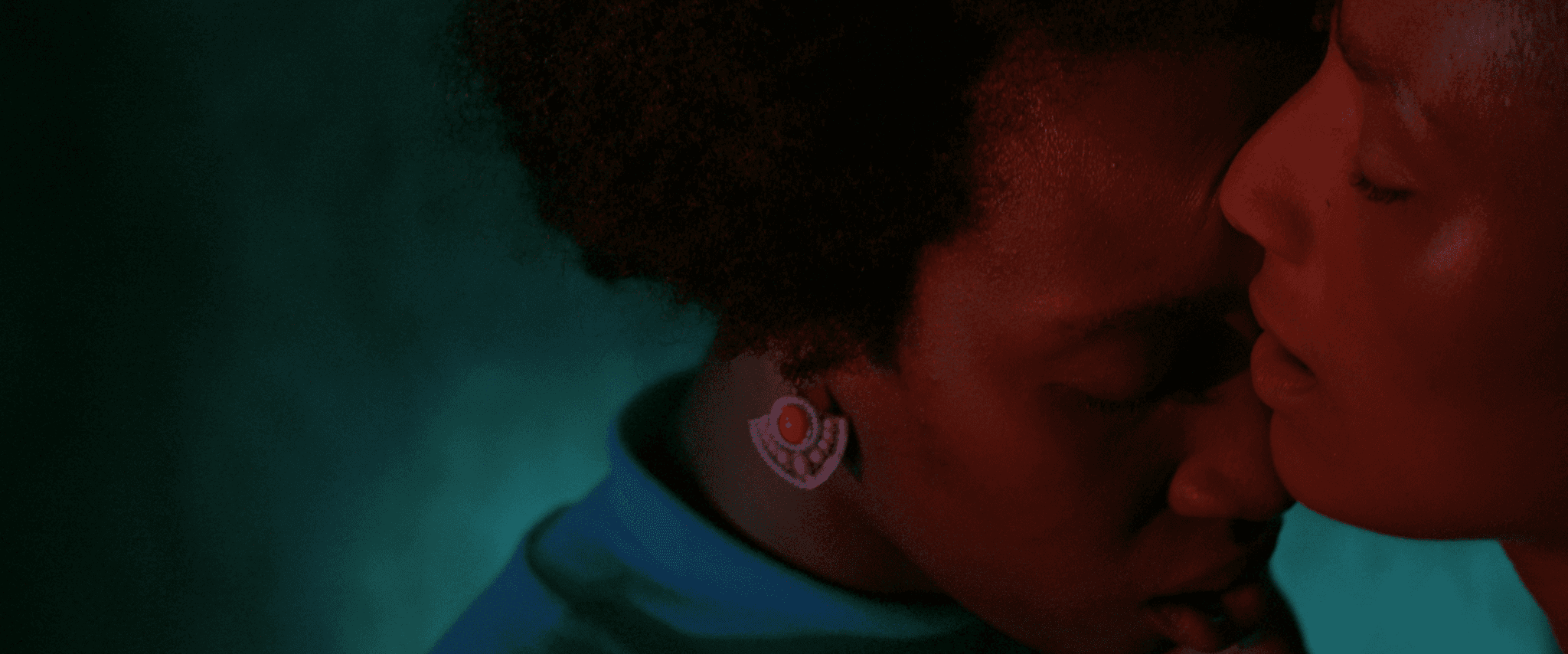 Still from the film "A felicidade delas" shows a close up of two people, standing so close they almost seem to be embracing, their faces almost touching. One person wears a blue turtleneck sweater, complementing the teal background and contrasting the warm red light on their faces.