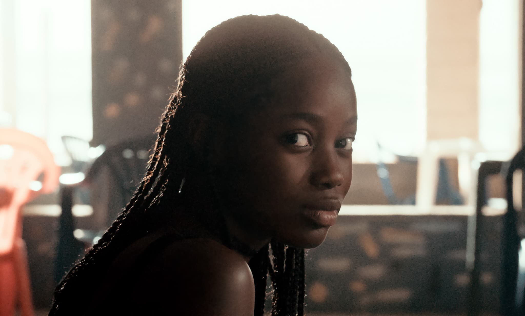 Still from the film Atlantics shows a young woman with long braids. She looks off screen as the light from a set of windows behind her illuminate her face.