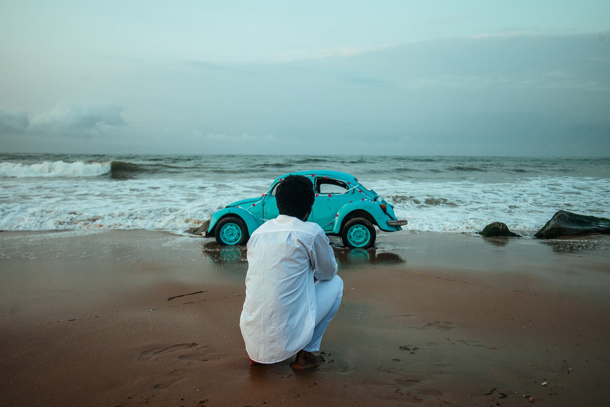 Still from the film "The Burial of Kojo" shows a burnt out car sitting on a beach. A person crouches in front of it as the waves roll on the beach in front of them.