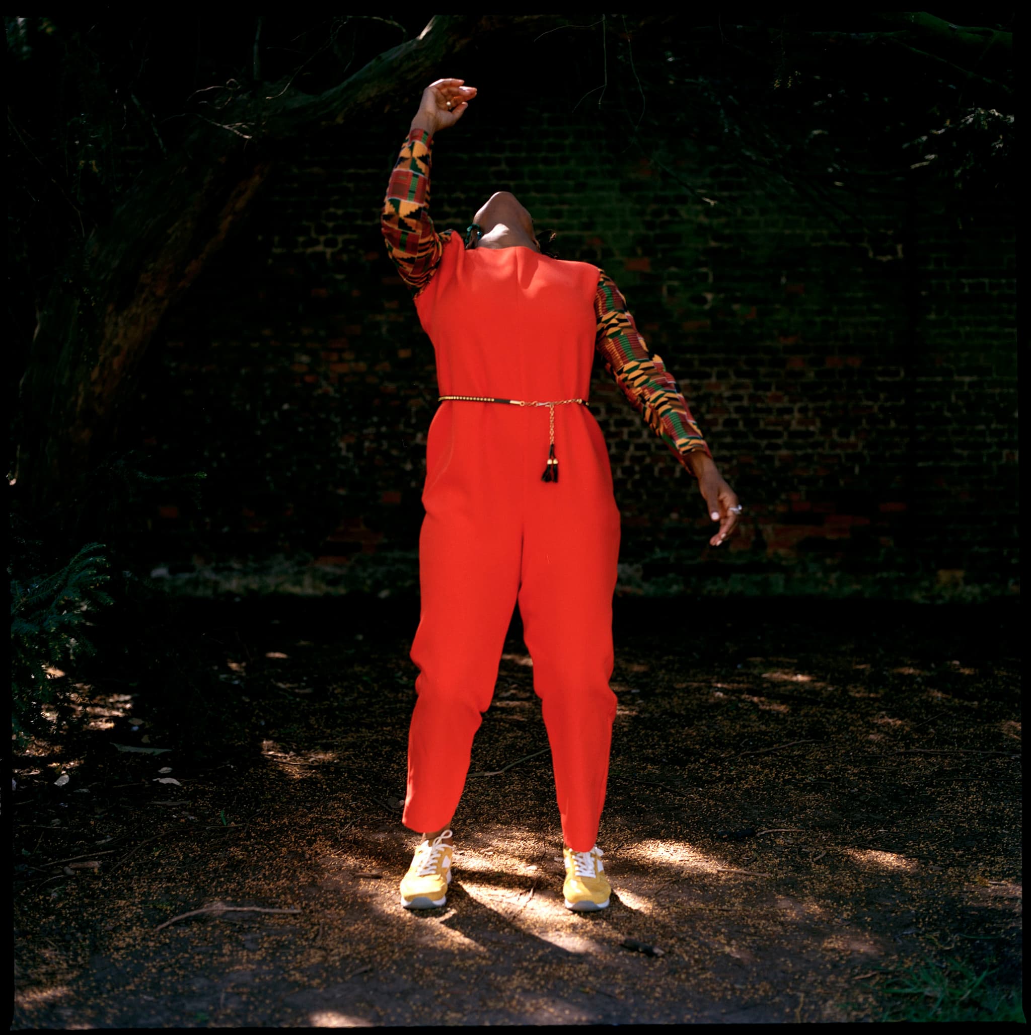 A person stands outside in front of a brick wall. They are wearing a red jumpsuit with kente print accents. Their head is thrown back towards the sky while one arm is arched above them.
