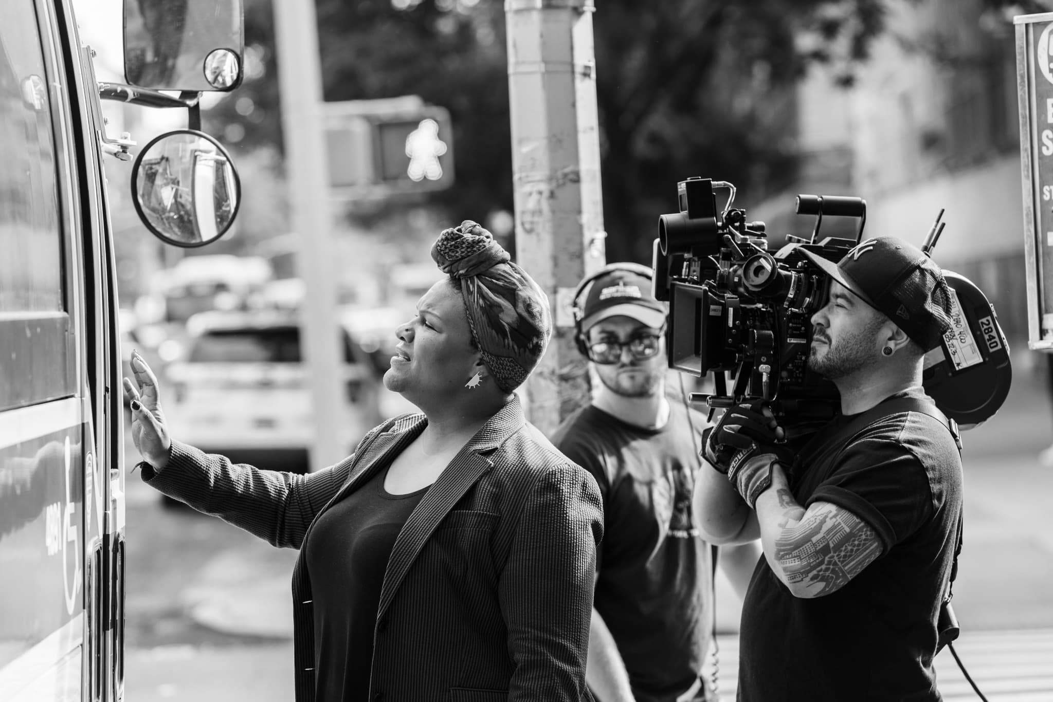 A black and white behind the scenes photo of a movie set shows a person wearing a headscarf and touching the window of a building. Behind them, a camera operator stands with a camera perched on their shoulder.