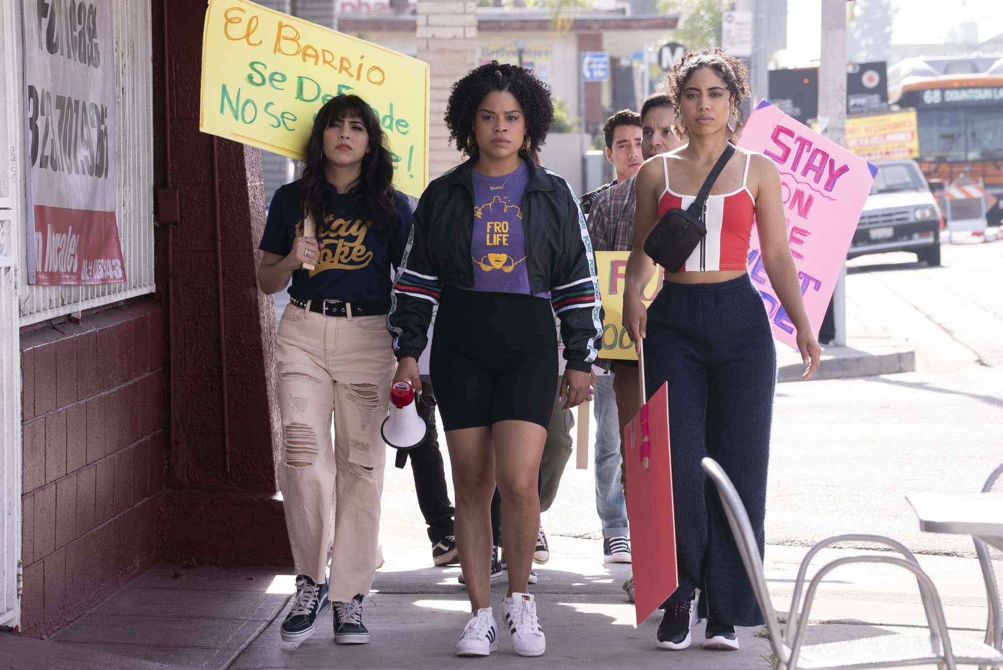Still from the show "Gentefied" shows a small group of people walking down a street. Several in the back of the group hold protest signs while one person in the front center holds a microphone.