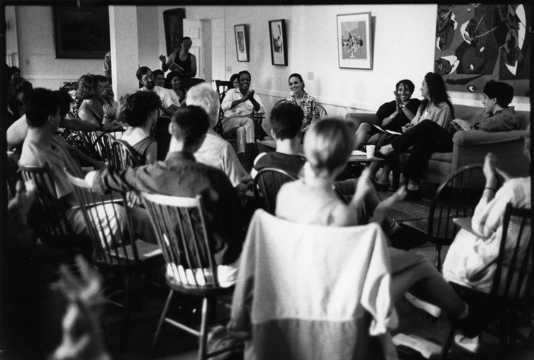 A group of people gather at a lecture. They sit in chairs and face a man, sitting and speaking at the front of the room.
