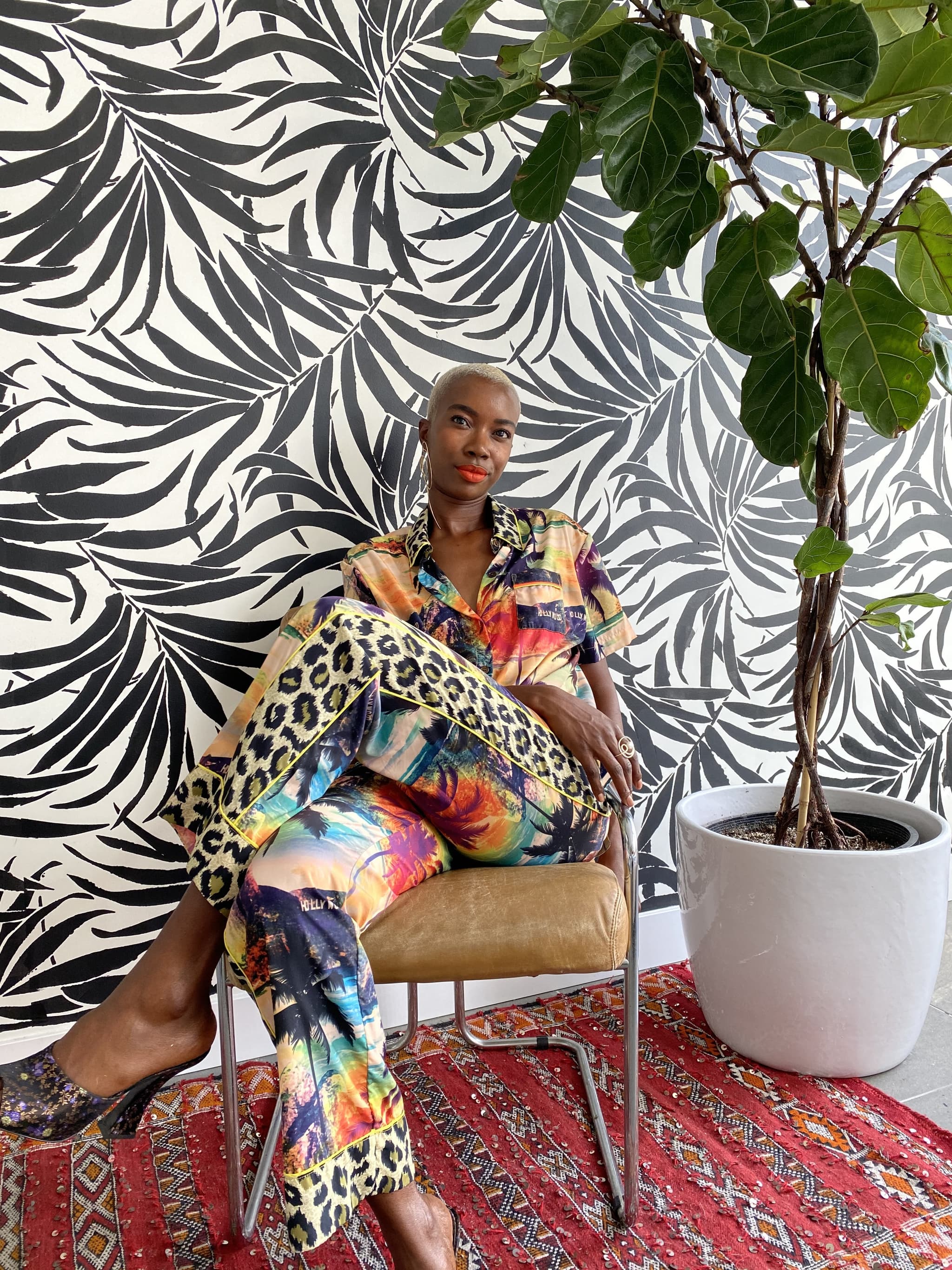 Photograph of Derica Cole Washington. The artist wears a multi-patterned jumpsuit and sits on a chair in front of a floral patterned wall. A tall houseplant sits next to them.