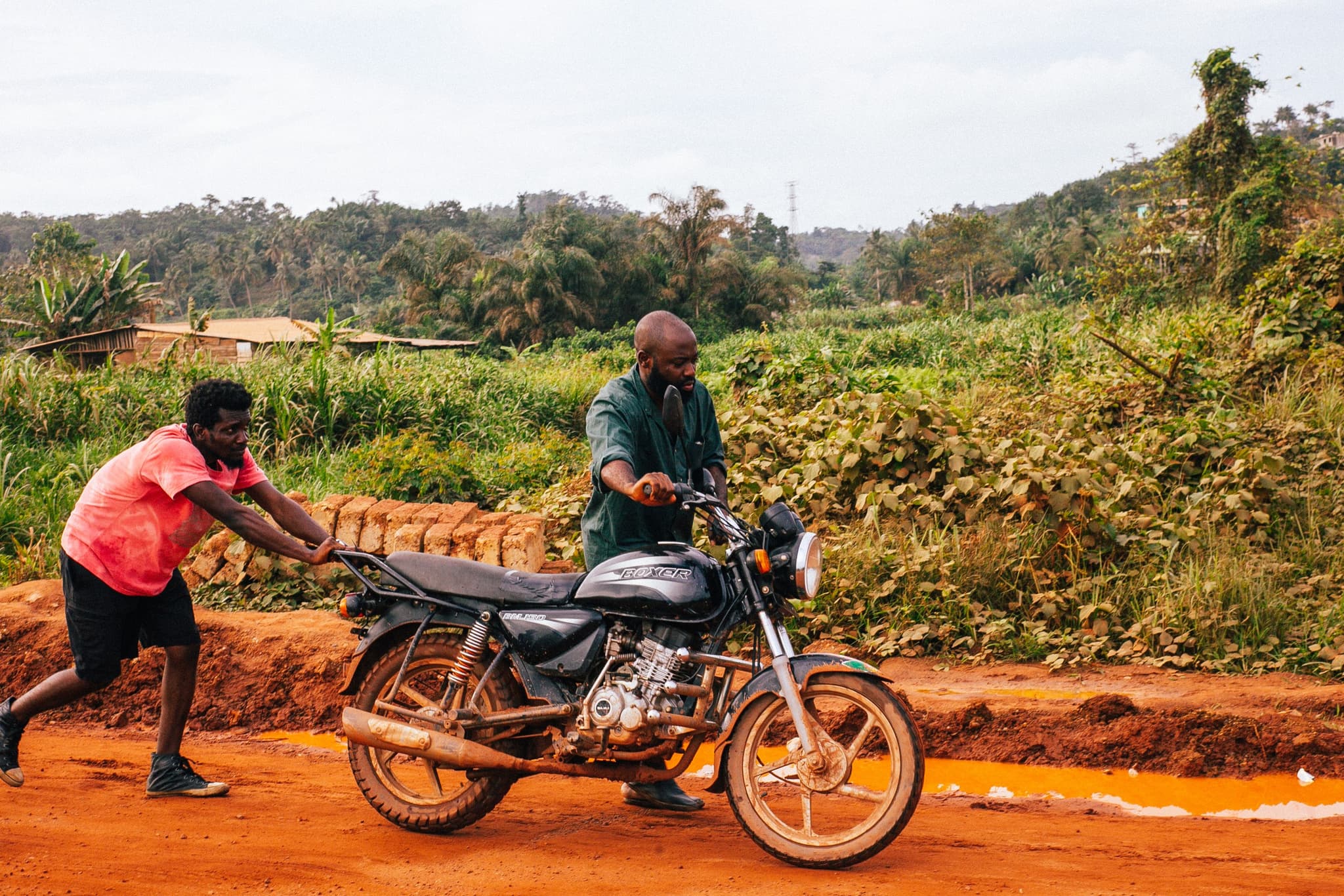 Still from the film "The Burial of Kojo" shows two men pushing a motorcycle on a red dirt road.