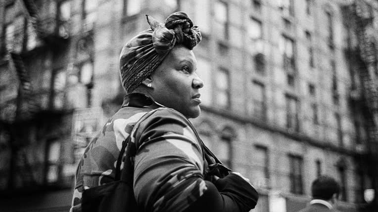 Still from "The Forty-Year-Old Version" is a black and white image of a person with a tied headscarf and large purse. They stand outside in front of a tall building ad look at something off-screen.
