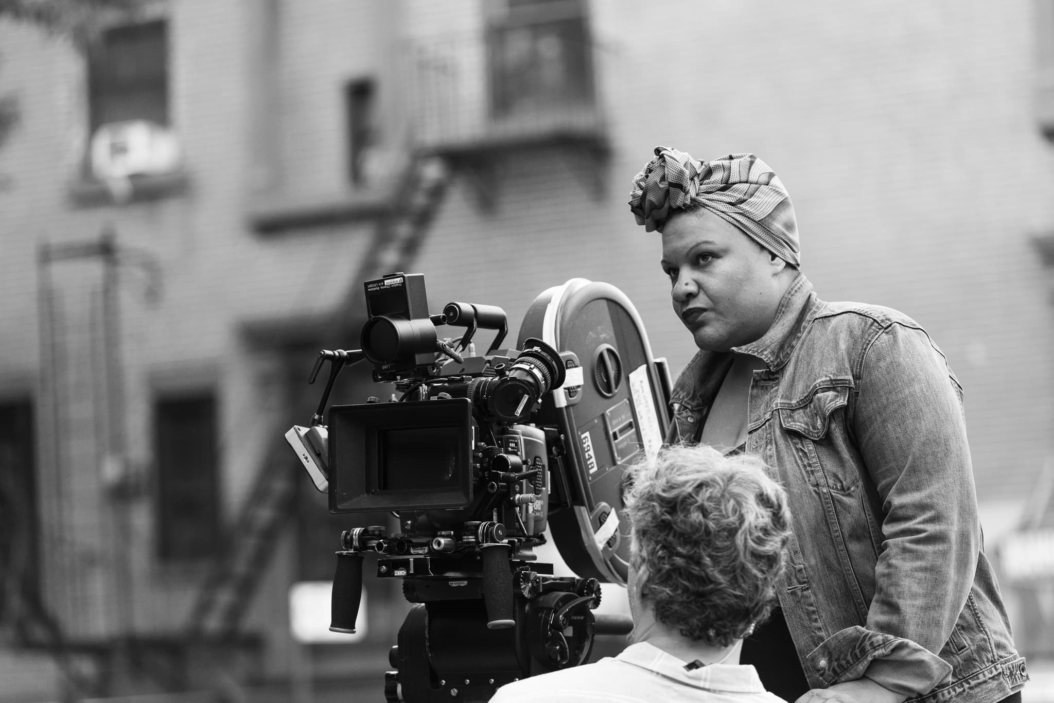 Image of a person standing in front of a large film camera on a film set. They wear a jean jacket and headscarf as they look at the camera.