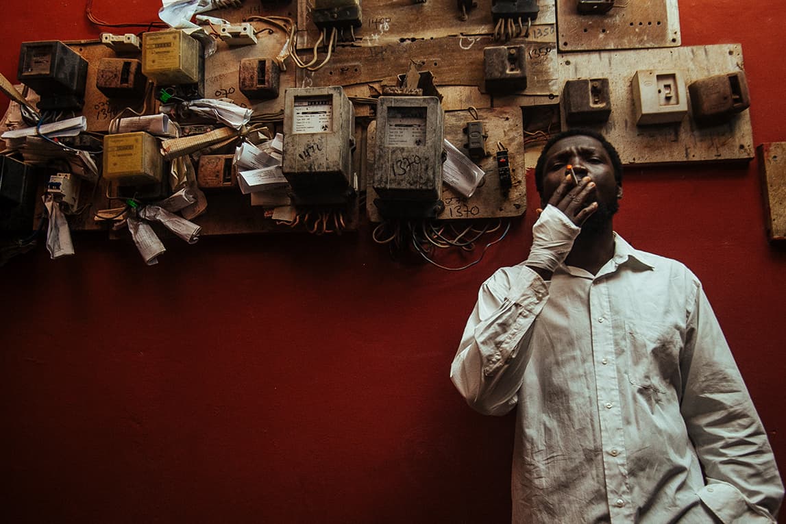 Still from the film "The Burial of Kojo" shoes a person wearing a button-down shirt and smoking a cigarette. Behind them, a wall filled with various rusted electrical components can be seen.
