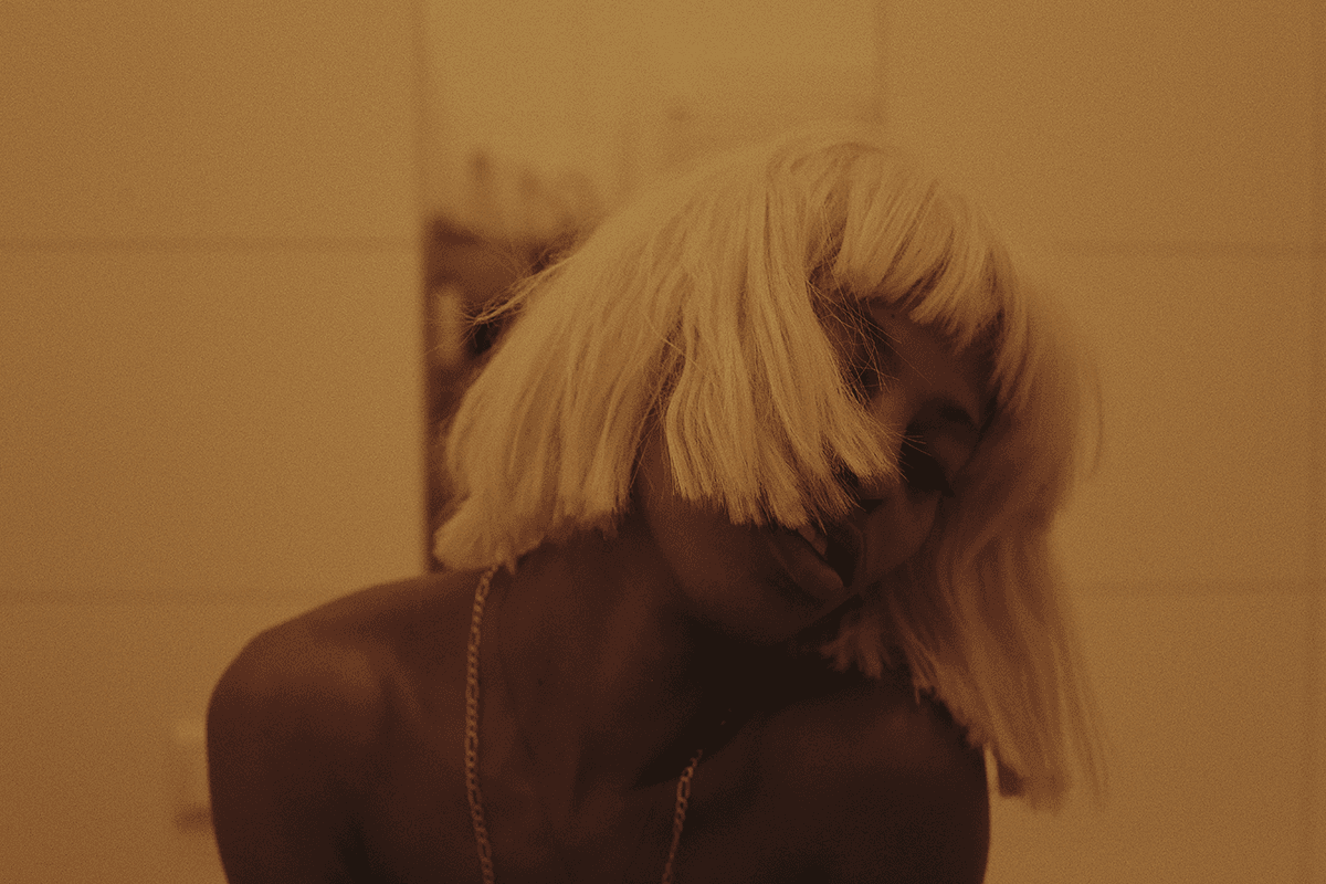 A still image from Rikki Wright's "A Song About Love" shows a Black person in a blonde wig, seemingly in a state of ecstasy or mid-dance, their head tilted to the side and hair obscuring much of their face.