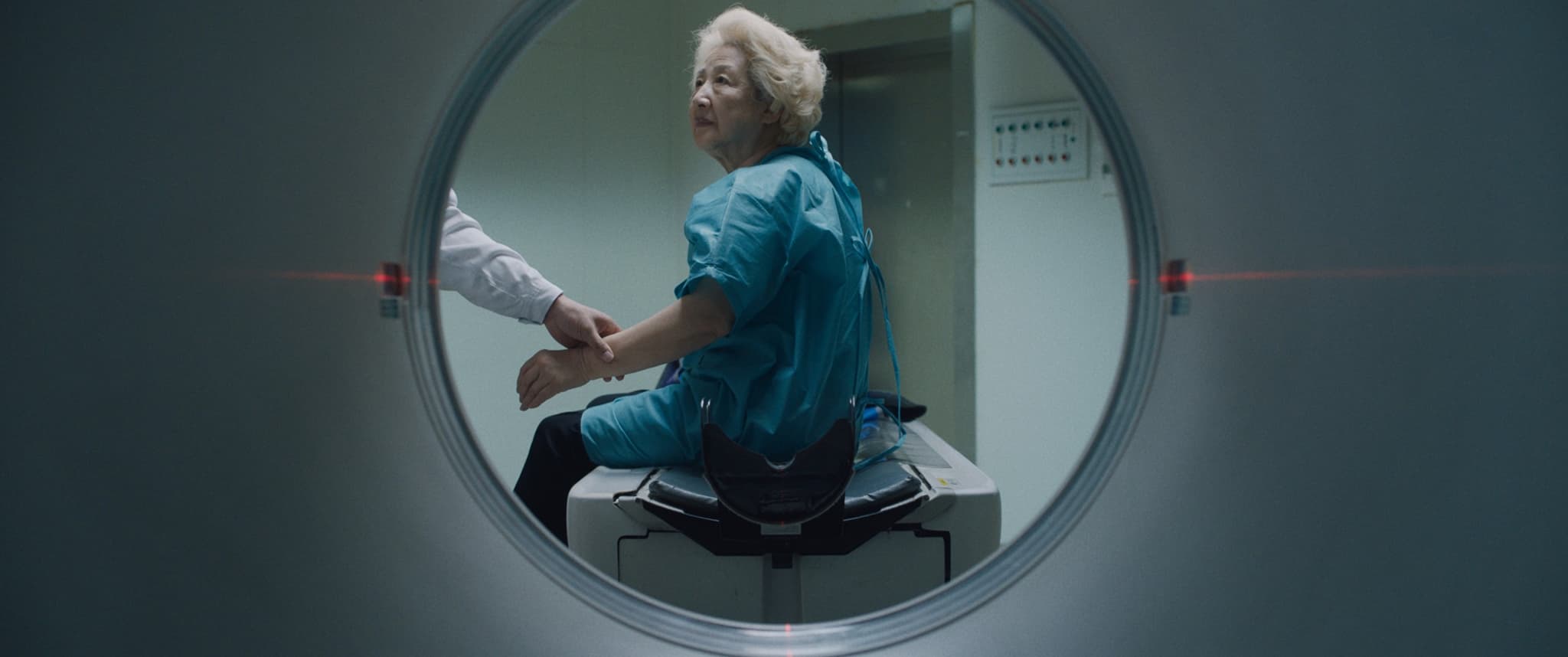 Still from the film "The Farewell" is an image taken from the inside of an MRI machine. A circle in the center of the frame shows an elderly woman in a blue medical gown, sitting and preparing to enter the machine. A hand can be seen holding on to her wrist as she looks, presumably at a medical professional, off screen.