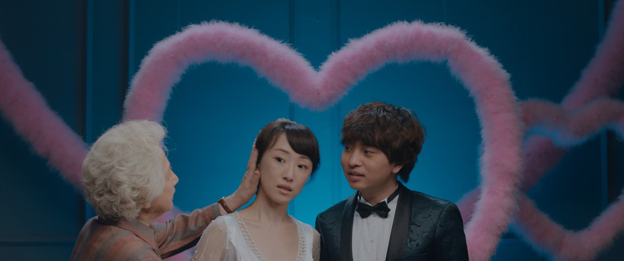 Still from the film "The Farewell" shows a couple preparing for a photo in front of a large, pink heart. An older woman leans in to touch the cheek of the young woman and fix her hair.