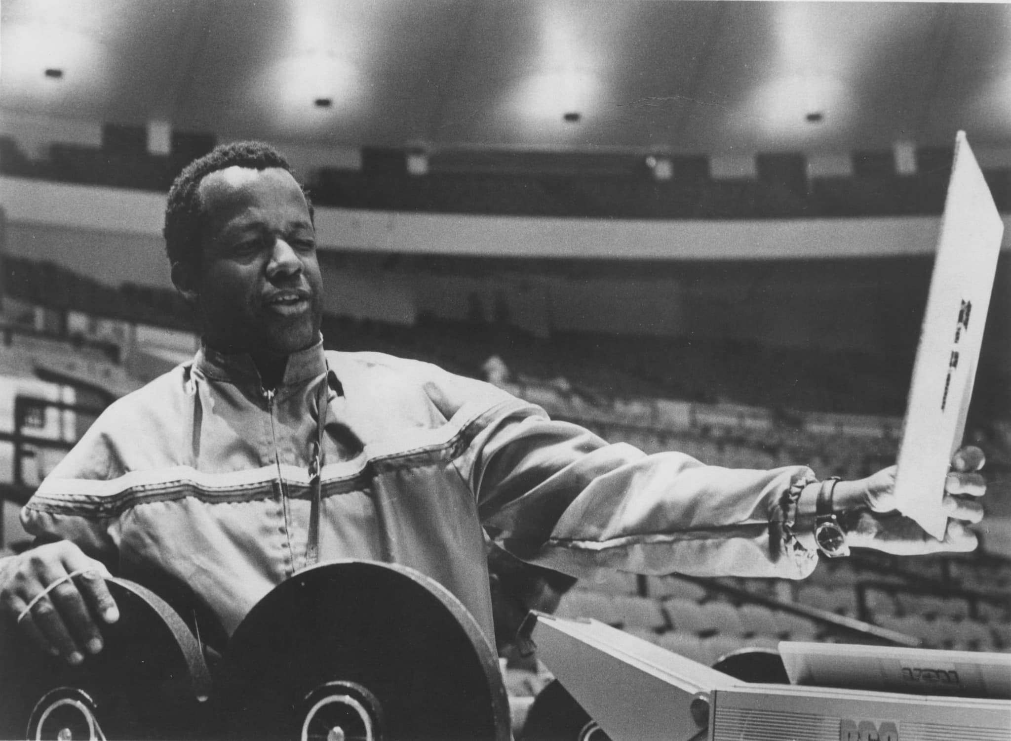 A Black Man stands in an auditorium. He holds out a piece of paper at arm's length in front of him and looks at it.