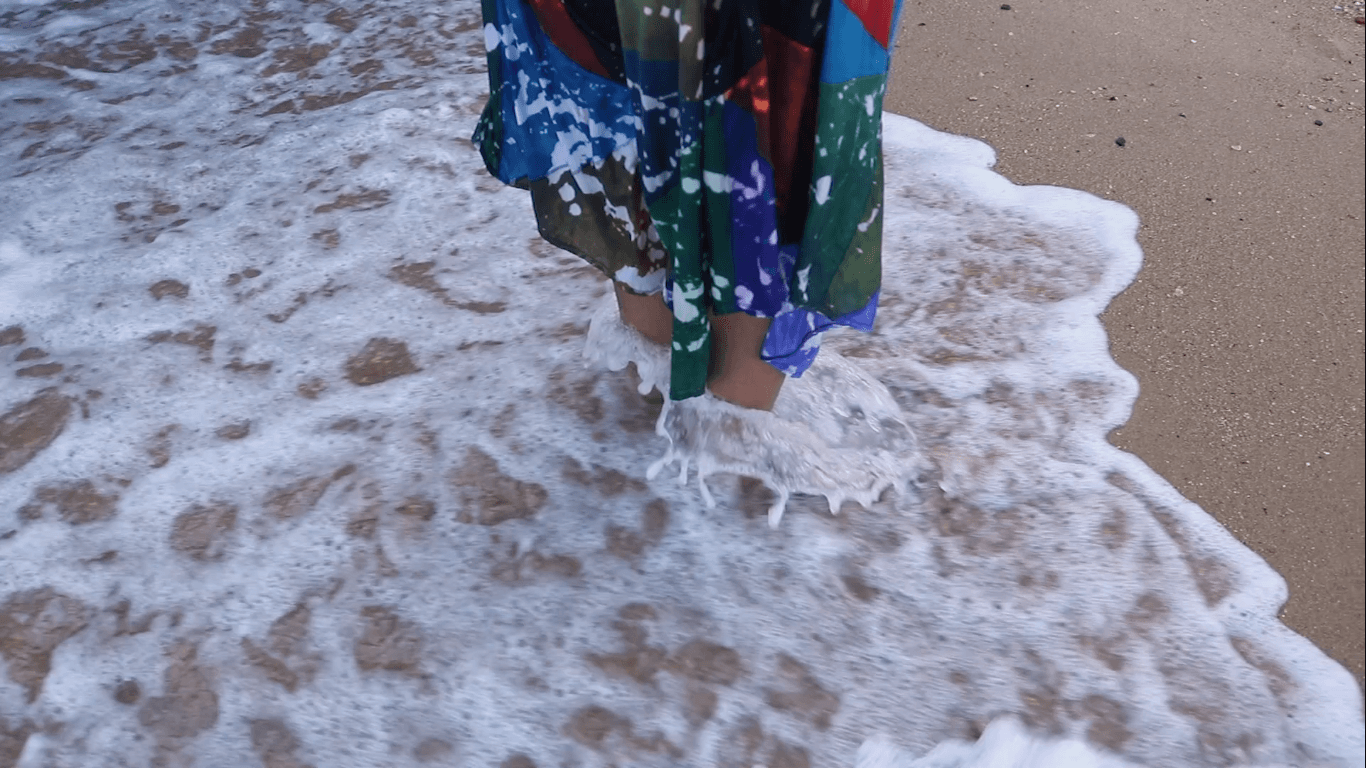 Still from "À beira do planeta mainha soprou a gente" shows the legs of a person wearing a long multicolored skirt as ocean waves wash over them. Water and seafoam circle their ankles and splashes the hem of the skirt.