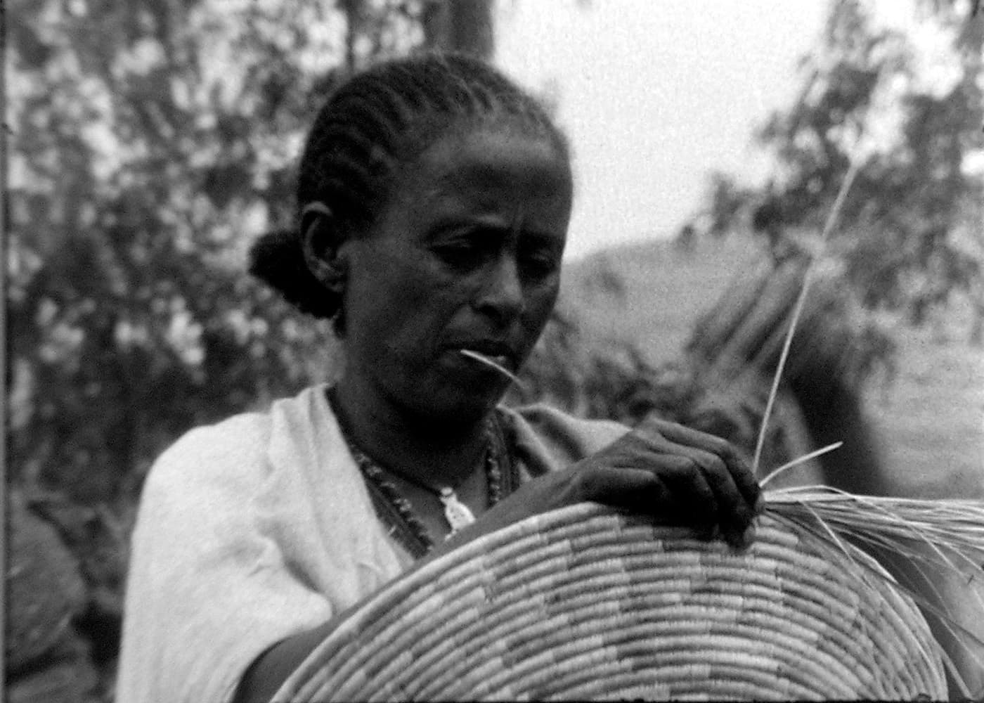 A Black person is sewing together a wicker basket. Photo in black and white.