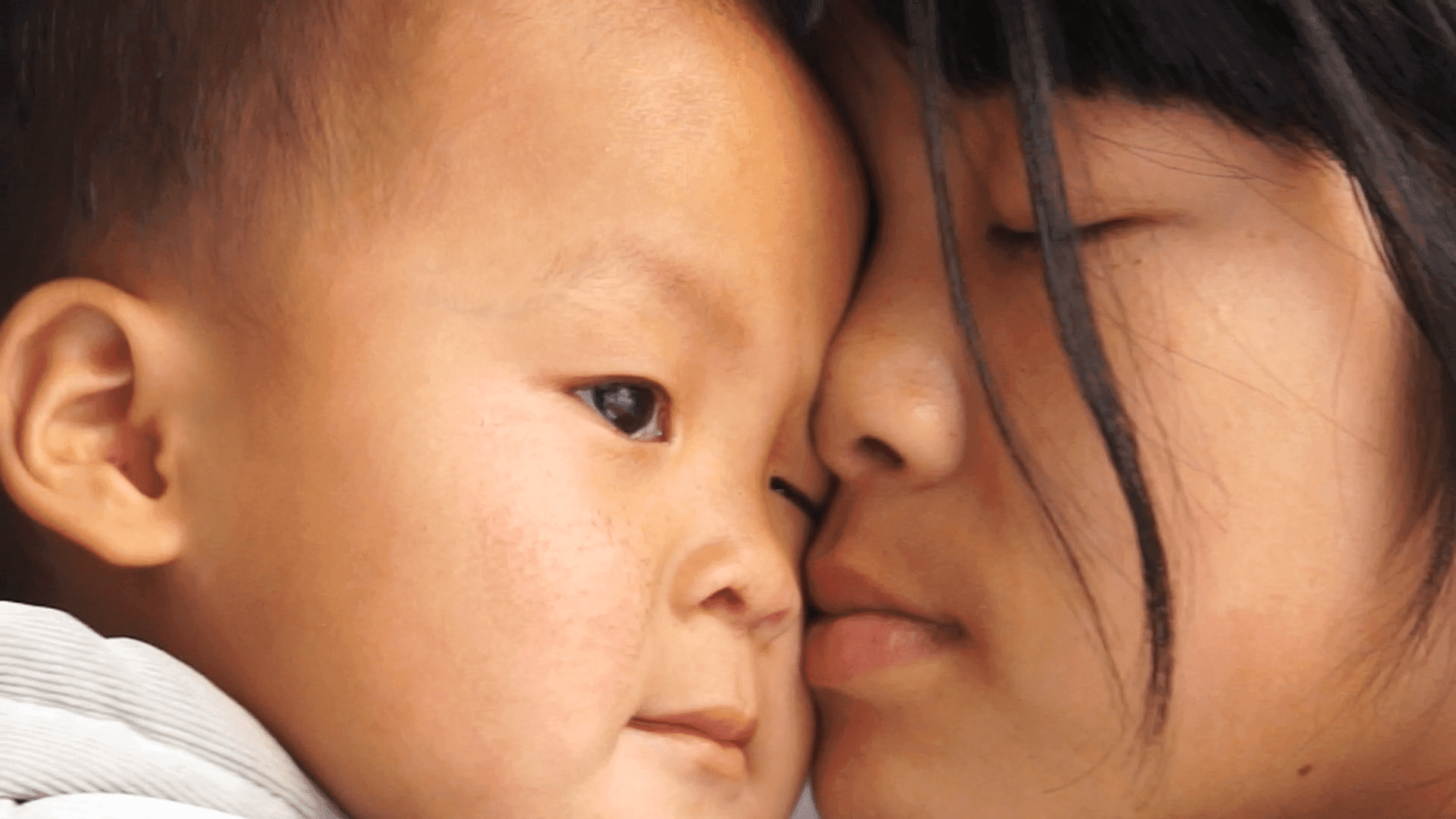 Two faces are pressed closely together, one is a baby and the other appears to be an adult. The adult has their eyes closed.