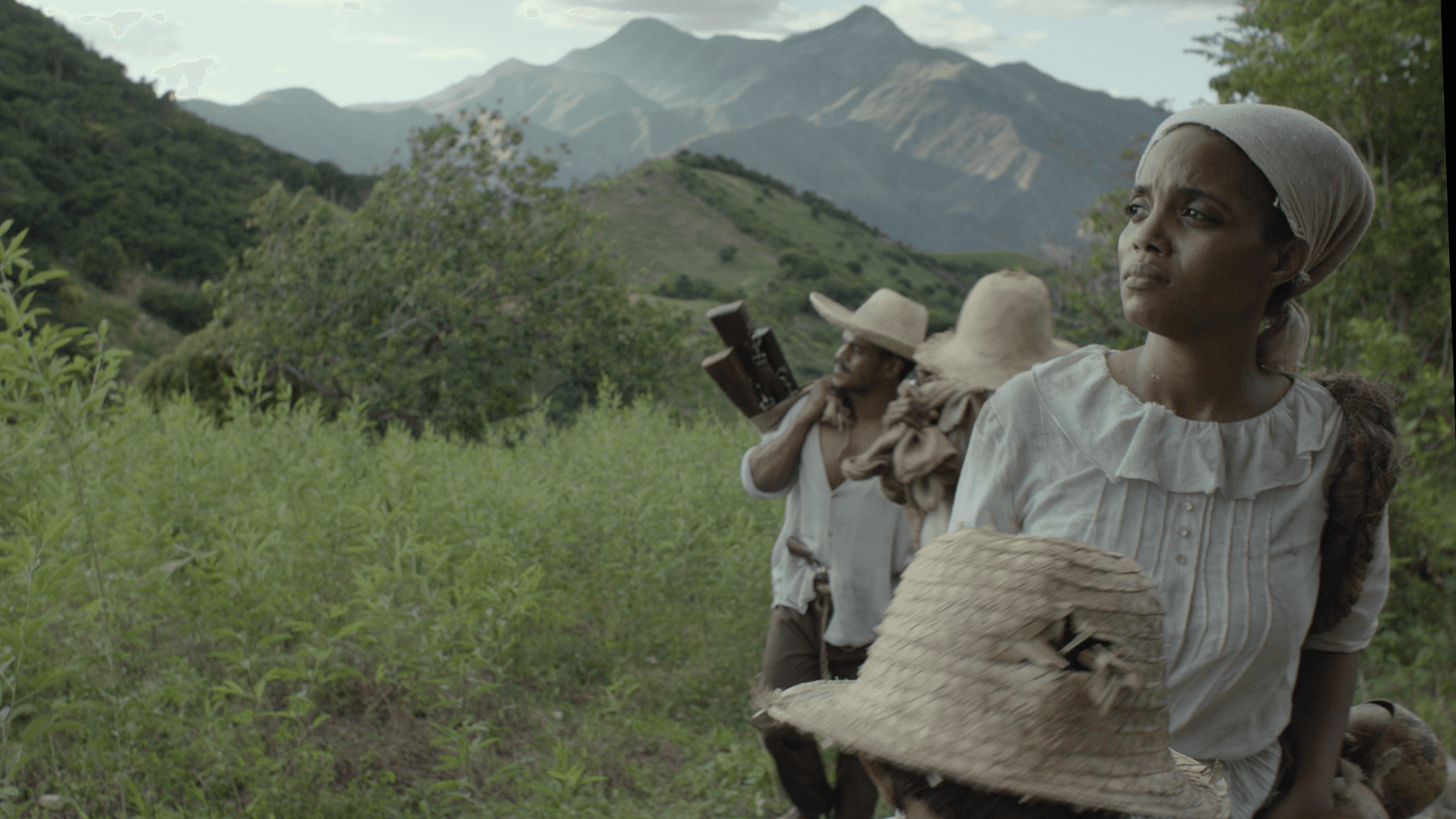 Image Description: A photo of a group of people, dressed in white and tan colored clothing, and walking in a mountainous area. The photo focuses on one woman, wearing a tan head covering and staring out into the distance. Behind the group, a series of mountains and vegetation can be seen.