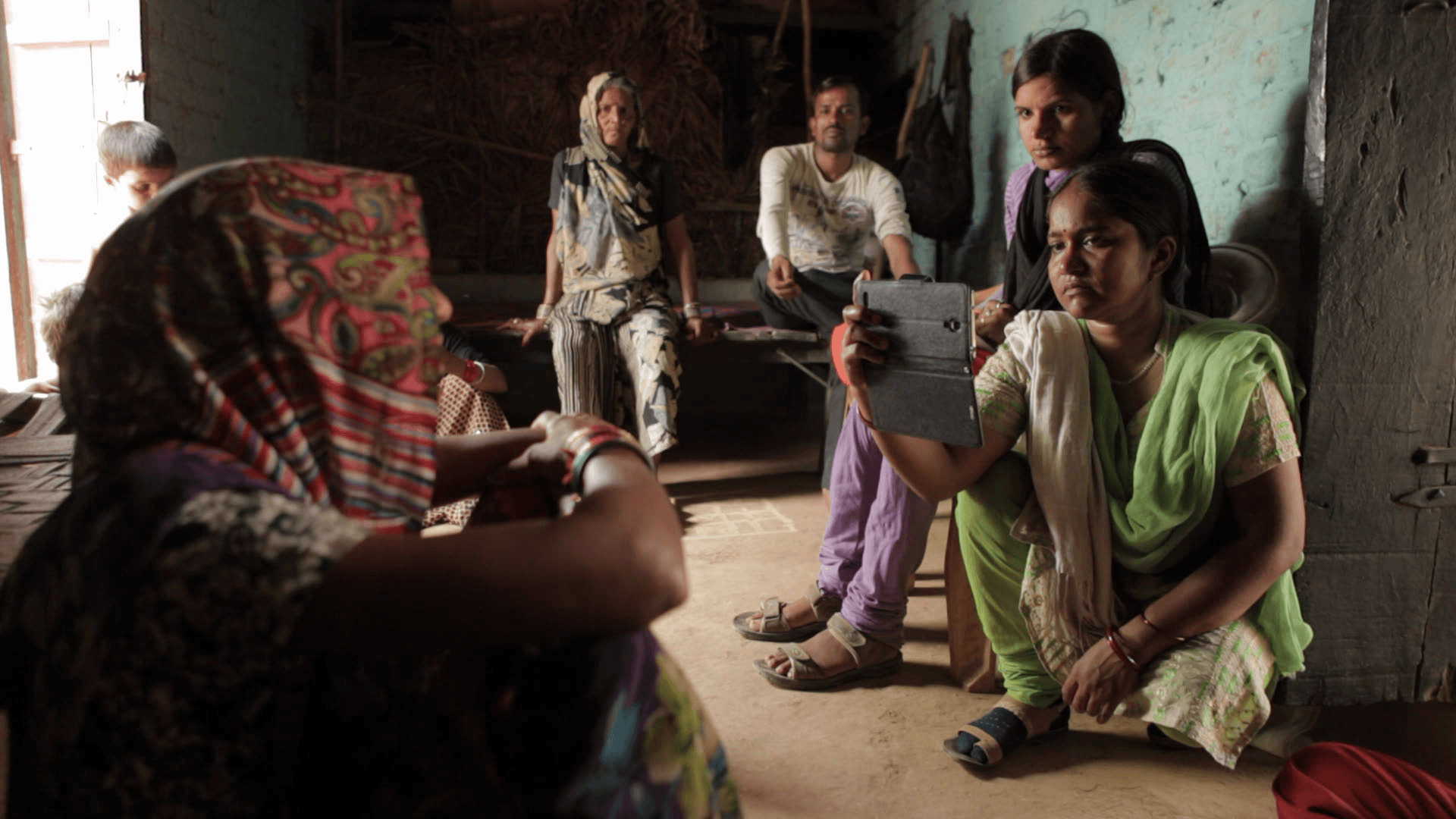 Still from the film "Writing With Fire" depicts a group of people sitting inside a house. One person holds up a smartphone to record an interview as the others look towards the individual speaking.