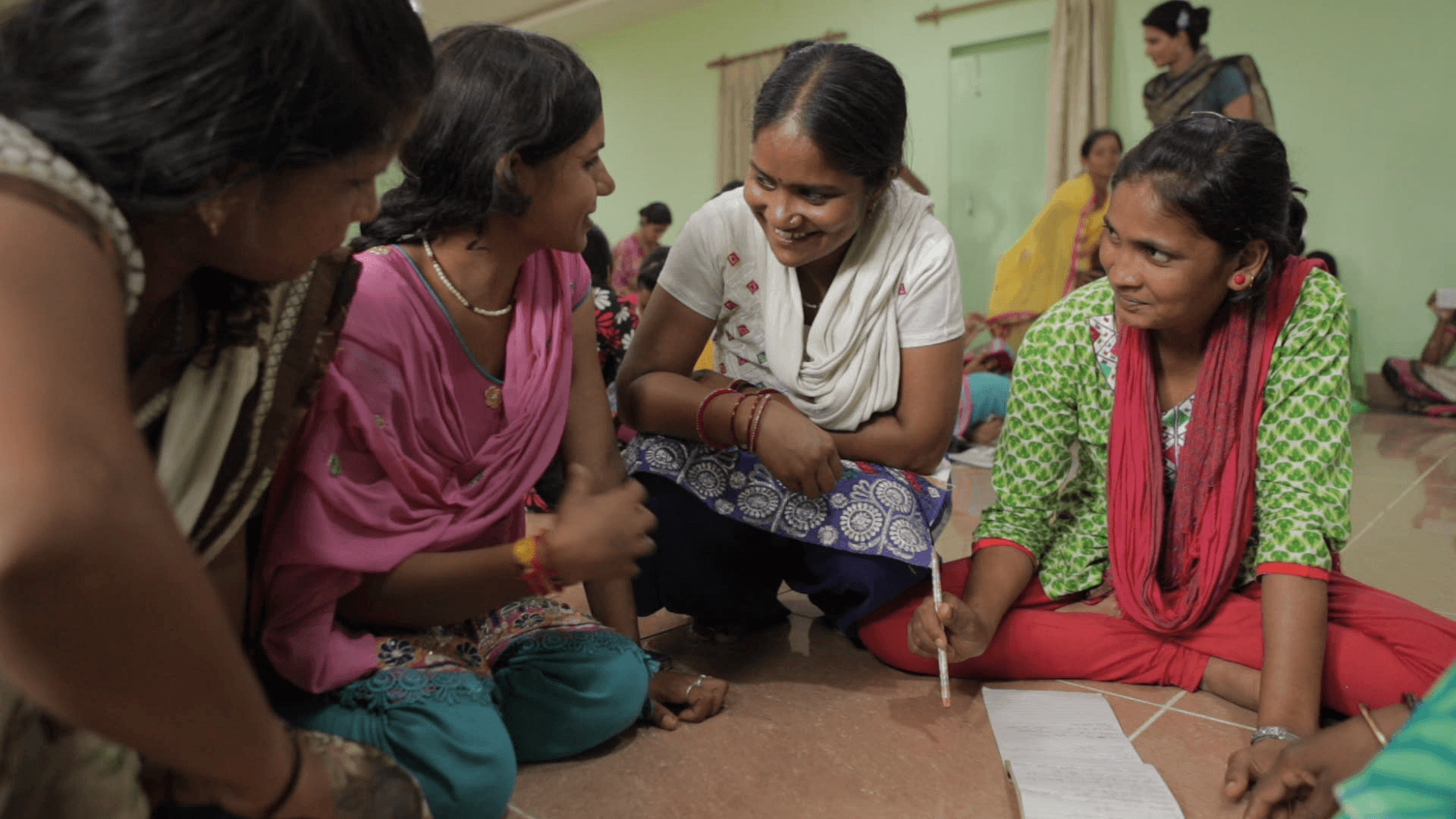 Still from the film "Writing With Fire" shows four women sitting on a floor and having a discussion. One woman holds a pencil and appears to be taking notes from their conversation on a notepad.