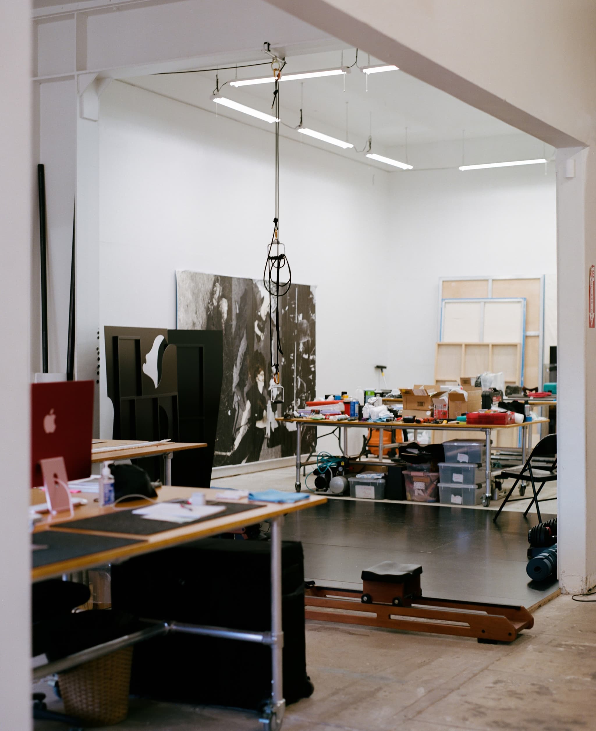 A photo by Pavielle Garcia shows the inside of Arthur Jafa's studio. There are a few large tables filled with ephemera and various art objects, a light hangs down from the tall ceiling between them and on the floor is an empty black space, perhaps its own canvas or space to create something else. On one of the walls is a very large poster, photo, or piece of art.