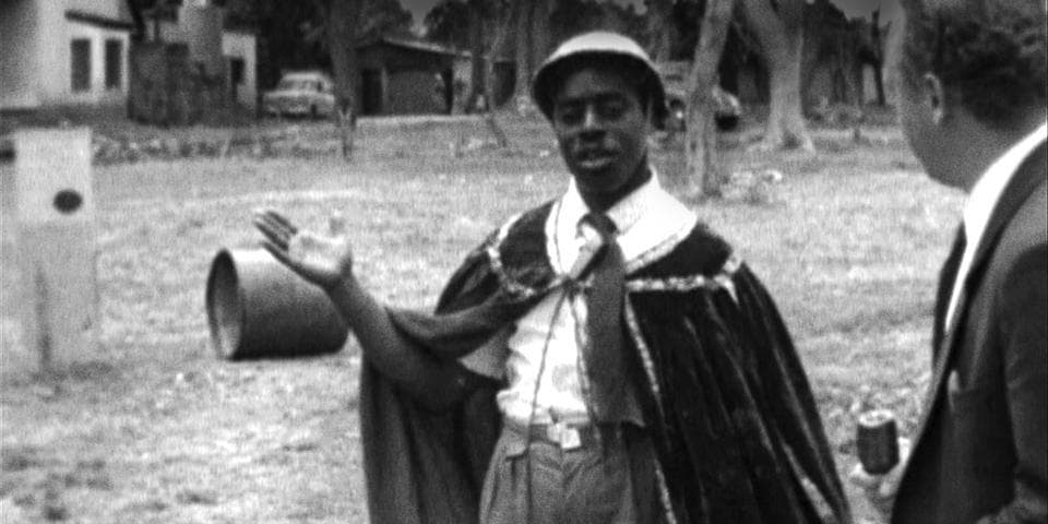 A Black person wears a cape and is showing something to a person off screen, in a black and white still