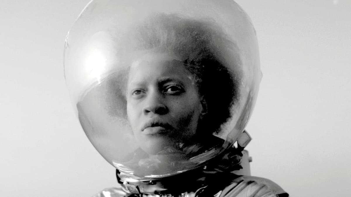 A Black person wears an astronaut suit (a clear globe around their head) in a black and white still.
