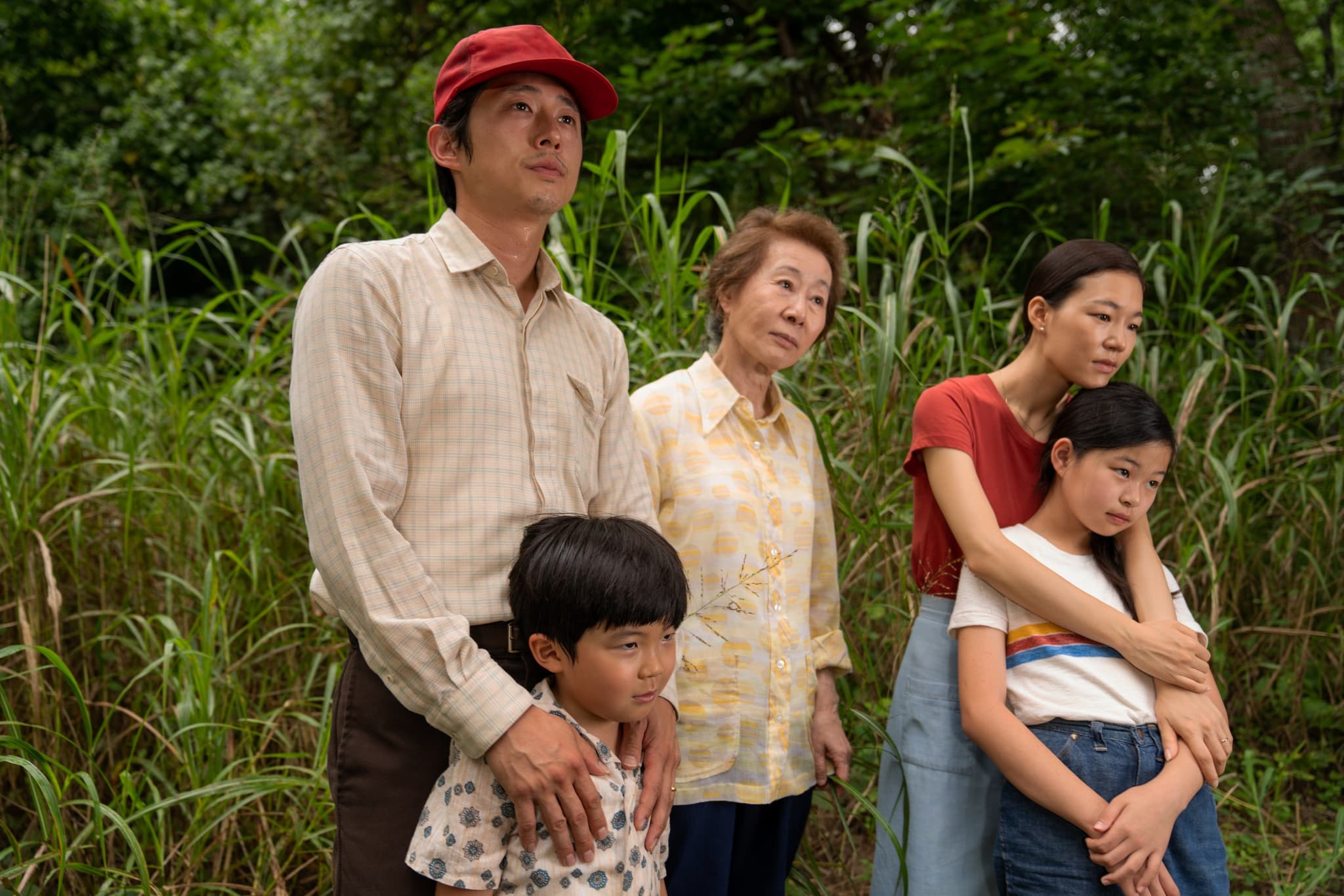 Still from the film "Minari" shows a Korean family of five, standing outside, embracing and looking off into the distance. The family consists of a father, mother, grandmother, and two young children (one boy and one girl). Behind them, tall green grass can be seen growing.
