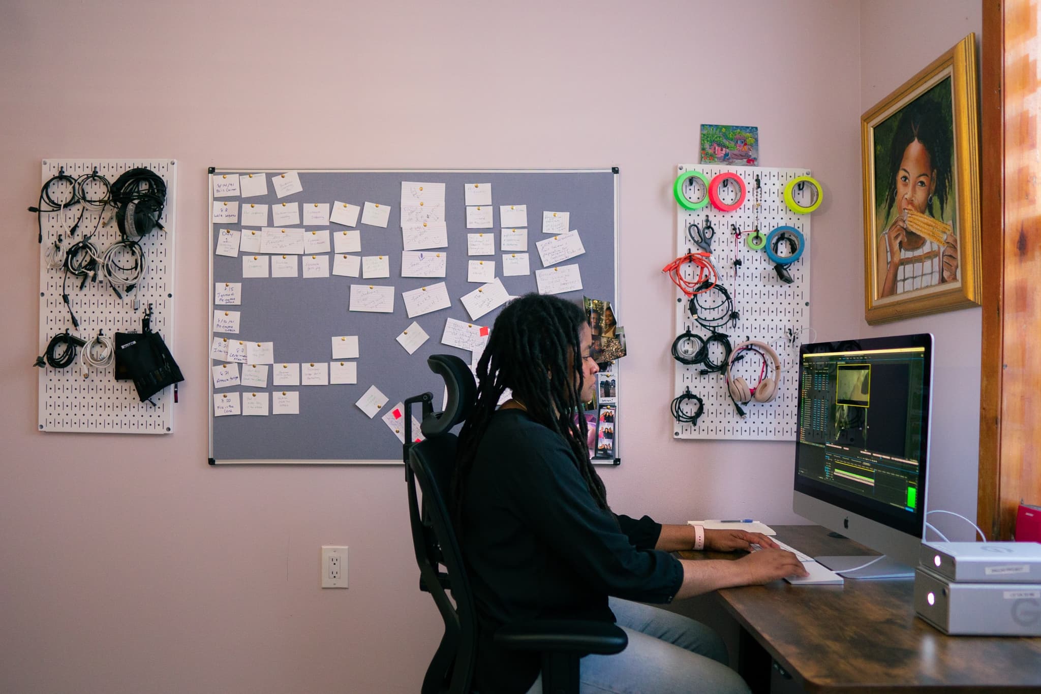 Image Description: A photo of editor Stefani Saintonge sitting at a computer desk. She looks at her desktop computer as her hands are positioned to type at a keyboard. Behind her, a bulletin board is filled with index cards and notes. On either side of it, various cords and different colored tape hang on white pegboards