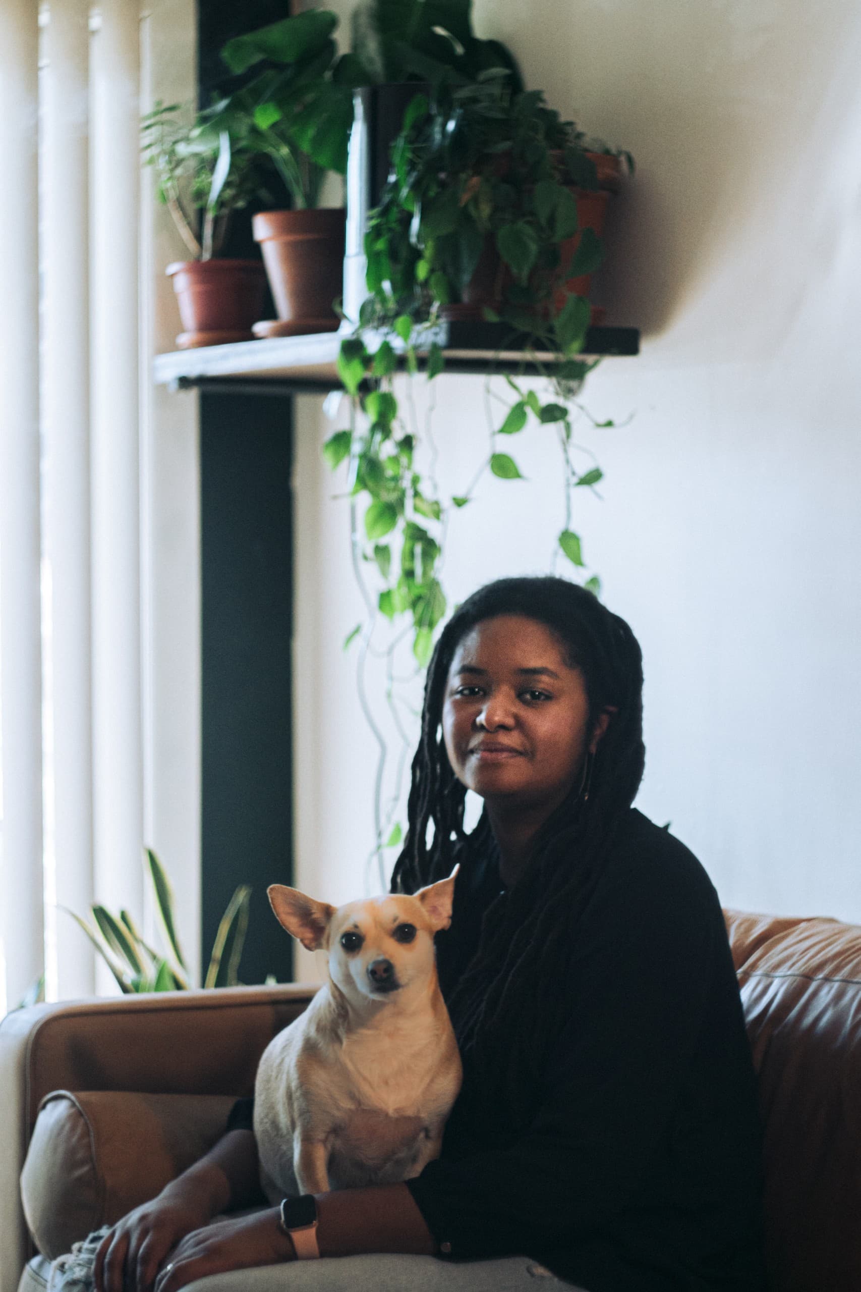 Image Description: A woman sits on a brown couch with a small dog in her lap. Behind her, a group of bright green plants can be seen on a shelf, contrasting with the warm muted colors in the remainder of the photo.