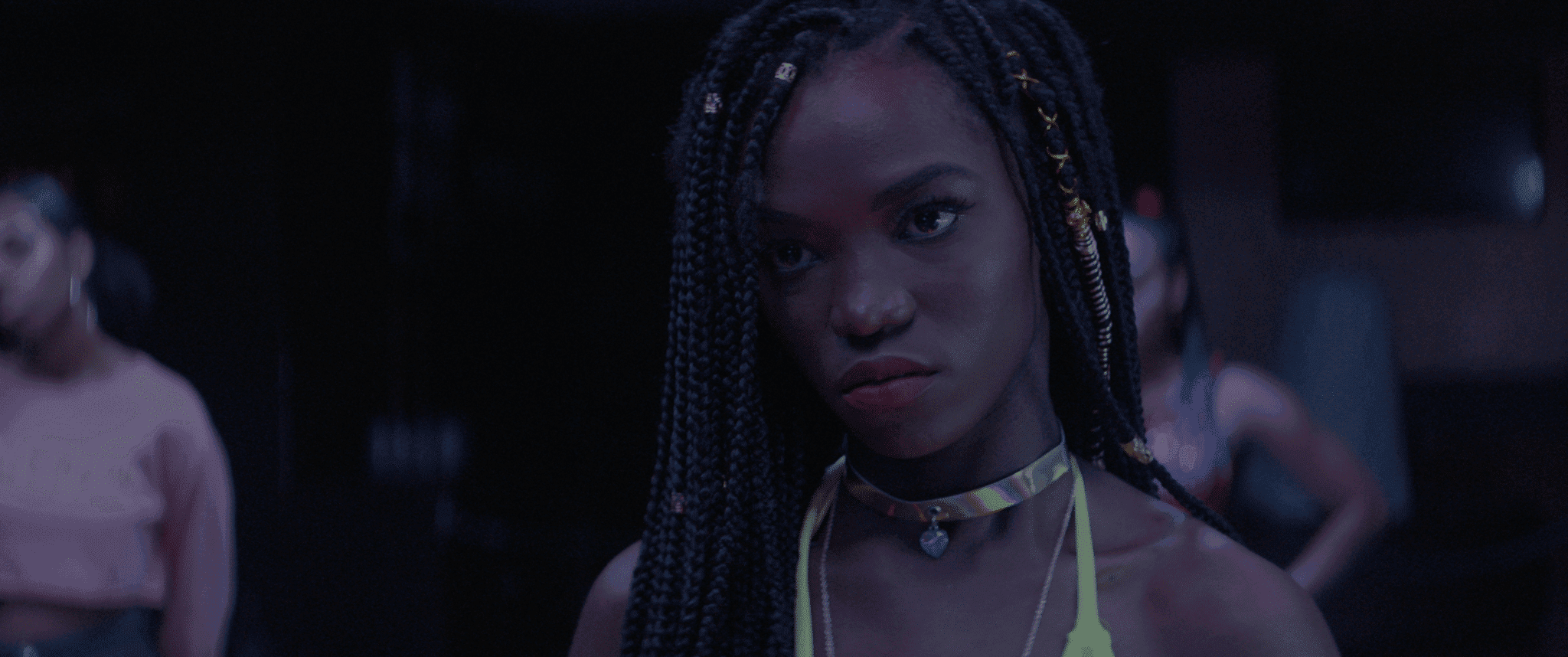 A Black person looks intently at someone off screen, they wear a gold necklace and their hair in braids, along with a yellow-neon bikini top.
