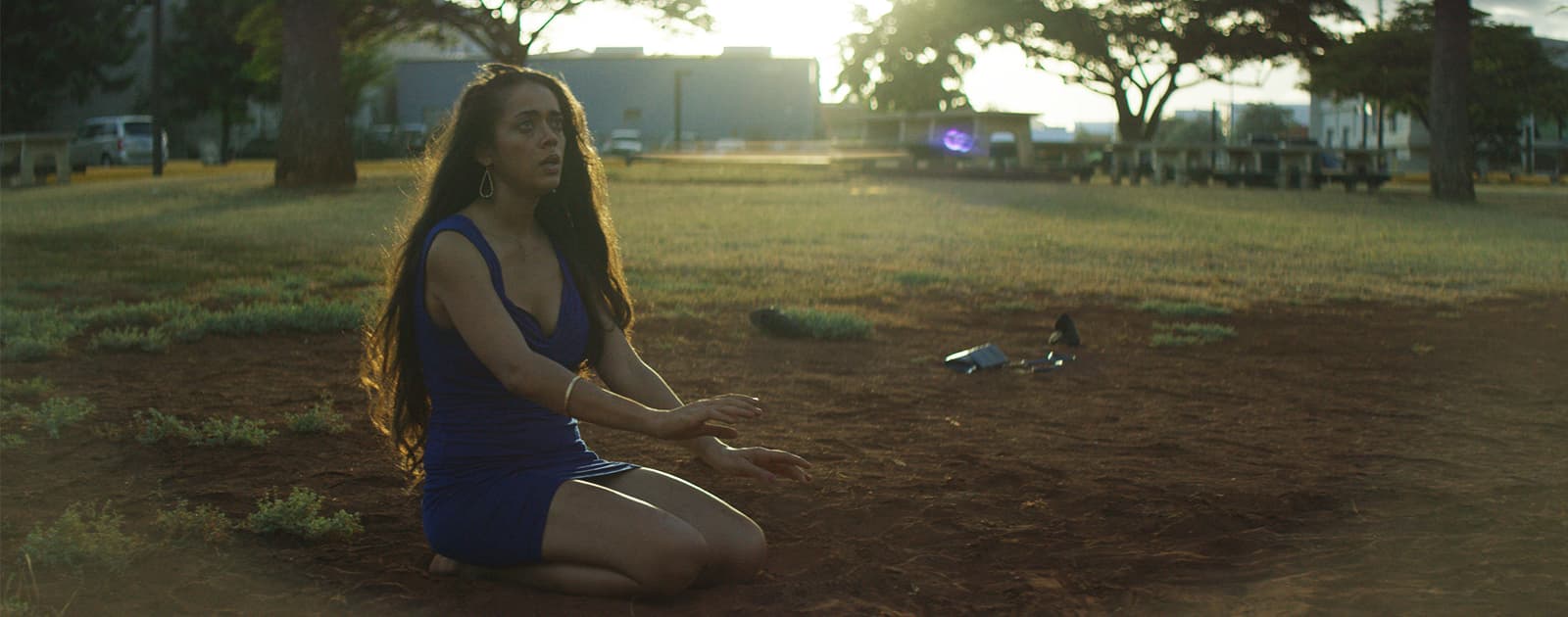 A Native Hawaiian person sits on the ground in the midday sunlight. They have their hands raised slightly, parallel to the ground.