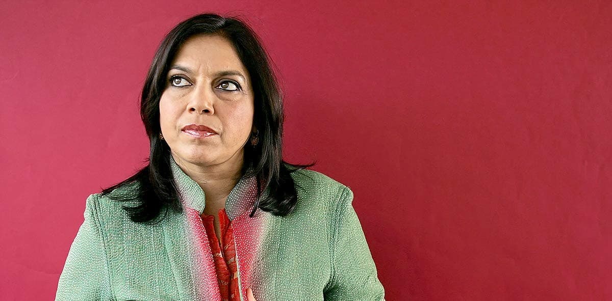 A headshot of Mira Nair, she is standing in front of a red wall. She is wearing a green and red coat and she is looking off to the side with a contemplative expression.