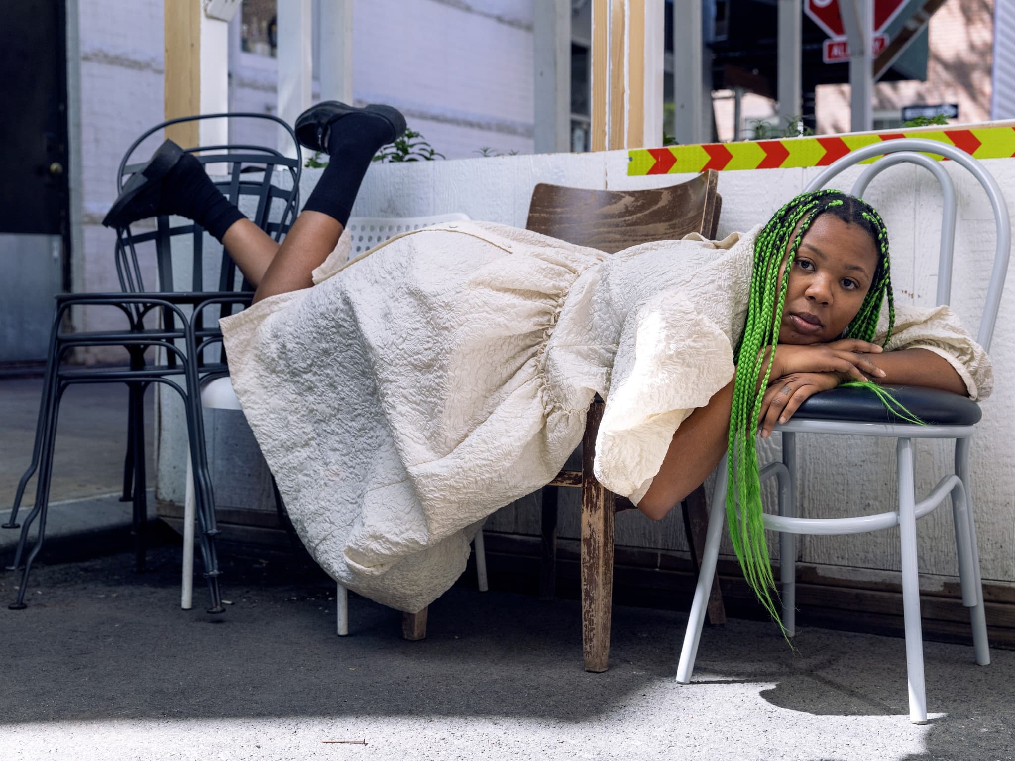 Martine Syms leans across two chairs, wearing a flowy cream colored dress and sporting bright green braids. They look at the camera directly.