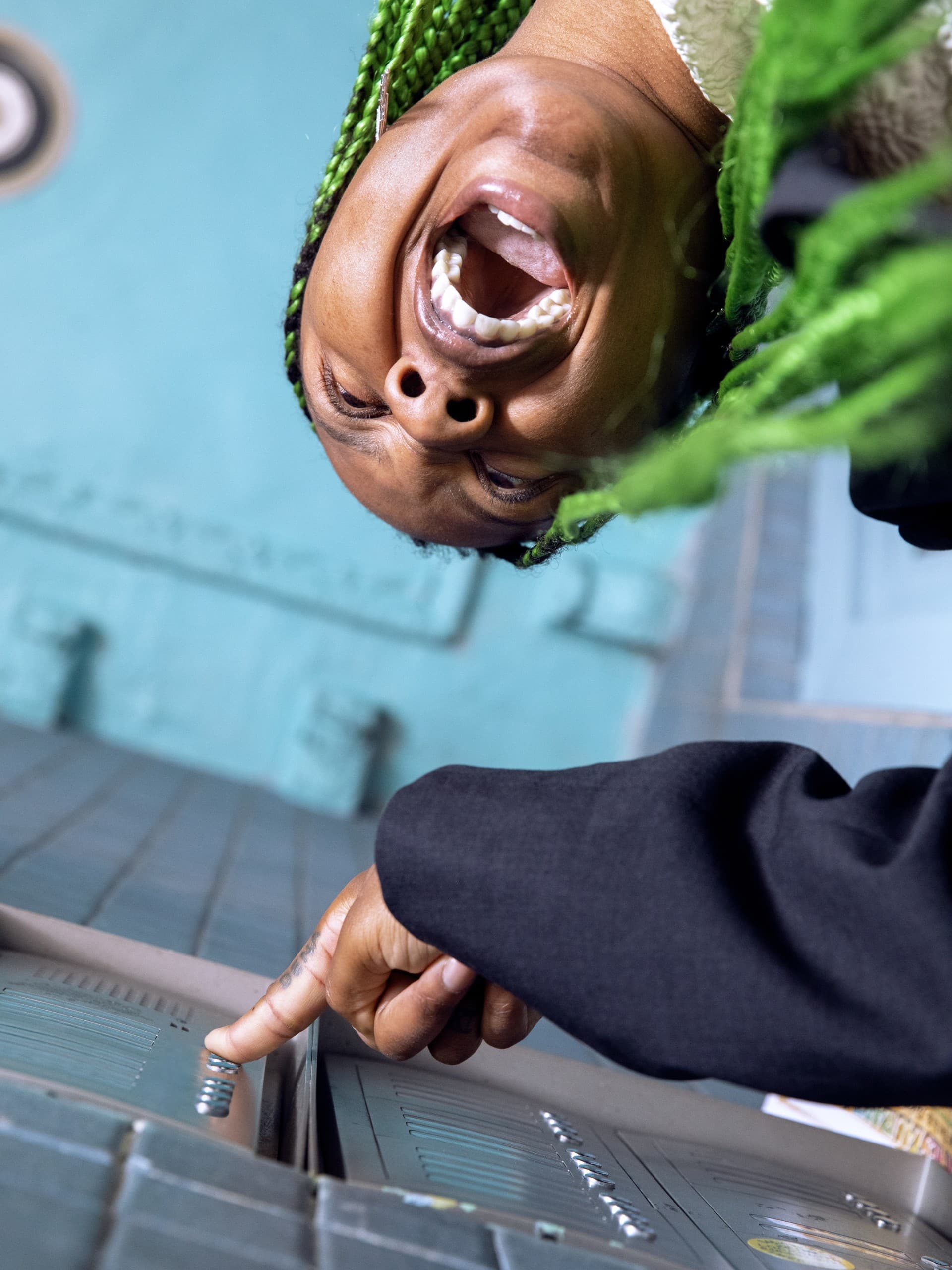 From beneath we look up at the image of Martine Syms pressing an apartment call button and apparently yelling. They have bright green braids and above them is a blue ceiling.