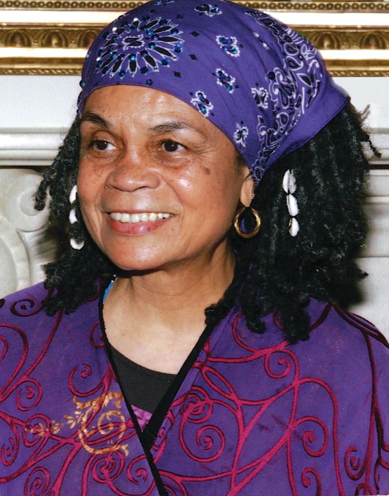 A headshot of Sonia Sanchez. She is wearing a purple suit jacket. Her hair is loc'd and pushed back with a purple bandana. She has cowrie shells in her hair and she is smiling.