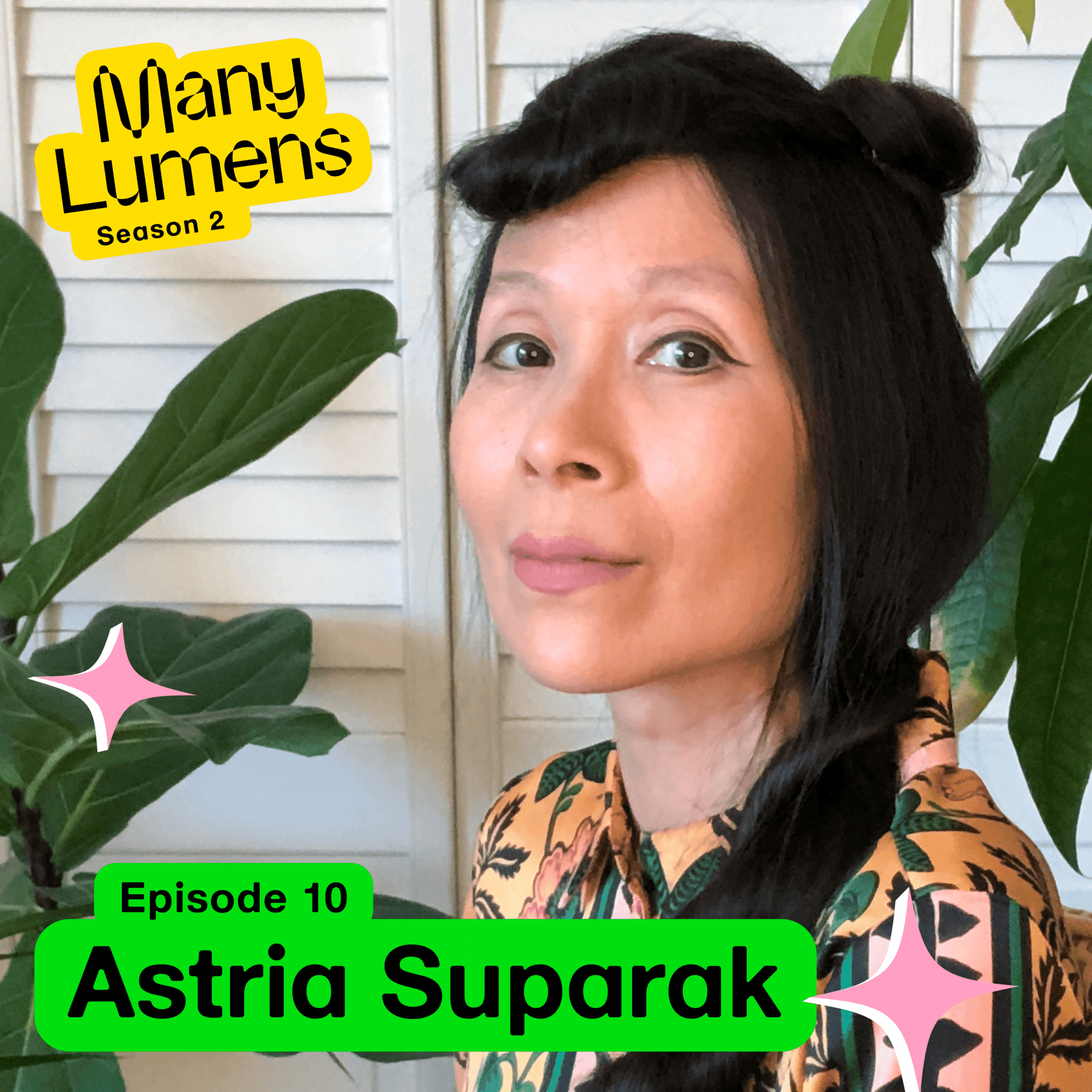 A photo of Astria Suparak. She is sitting in a wicker chair and there are plants behind her. She is wearing a gray skirt and a patterned top. She is smiling and looking at the camera. Many Lumens Season 2 Episode 10 guest.