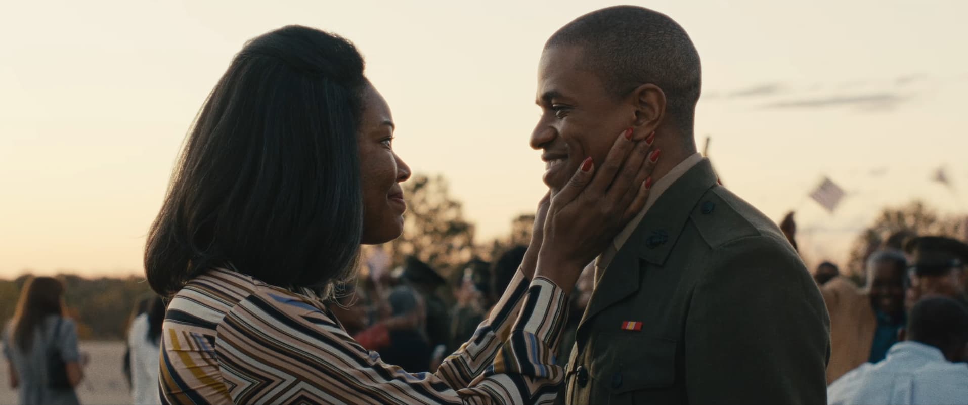 A Black man smiles at an older Black person who is cupping their face in their hands, looking lovingly at them - perhaps their mother. The man wears a military uniform but has their hat removed.