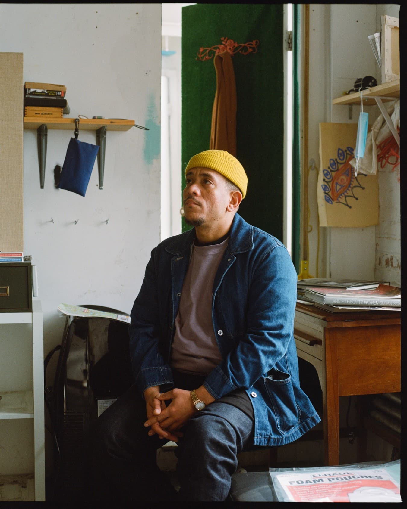 A photo of Kenny Rivero sitting in his studio. A brown skinned man sits in an art studio looking slightly upward.