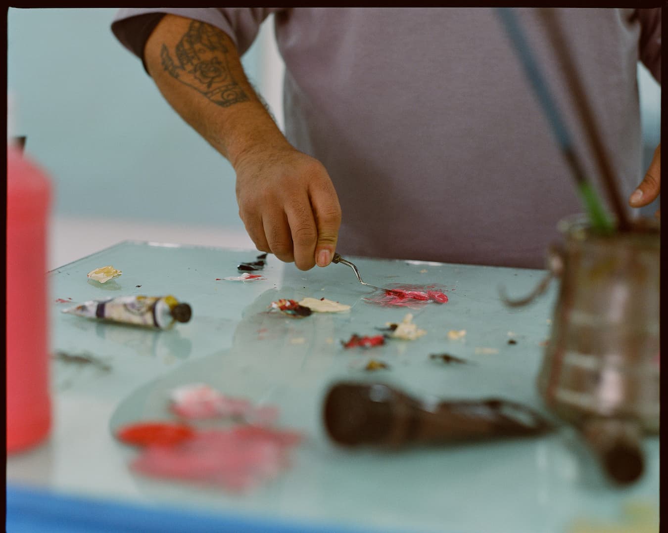 A photo of Kenny Rivero in studio painting on glass. A tattooed arm of a brown skinned man holds a palette knife, paint tubes scattered amongst globs of paint on a glass surface.