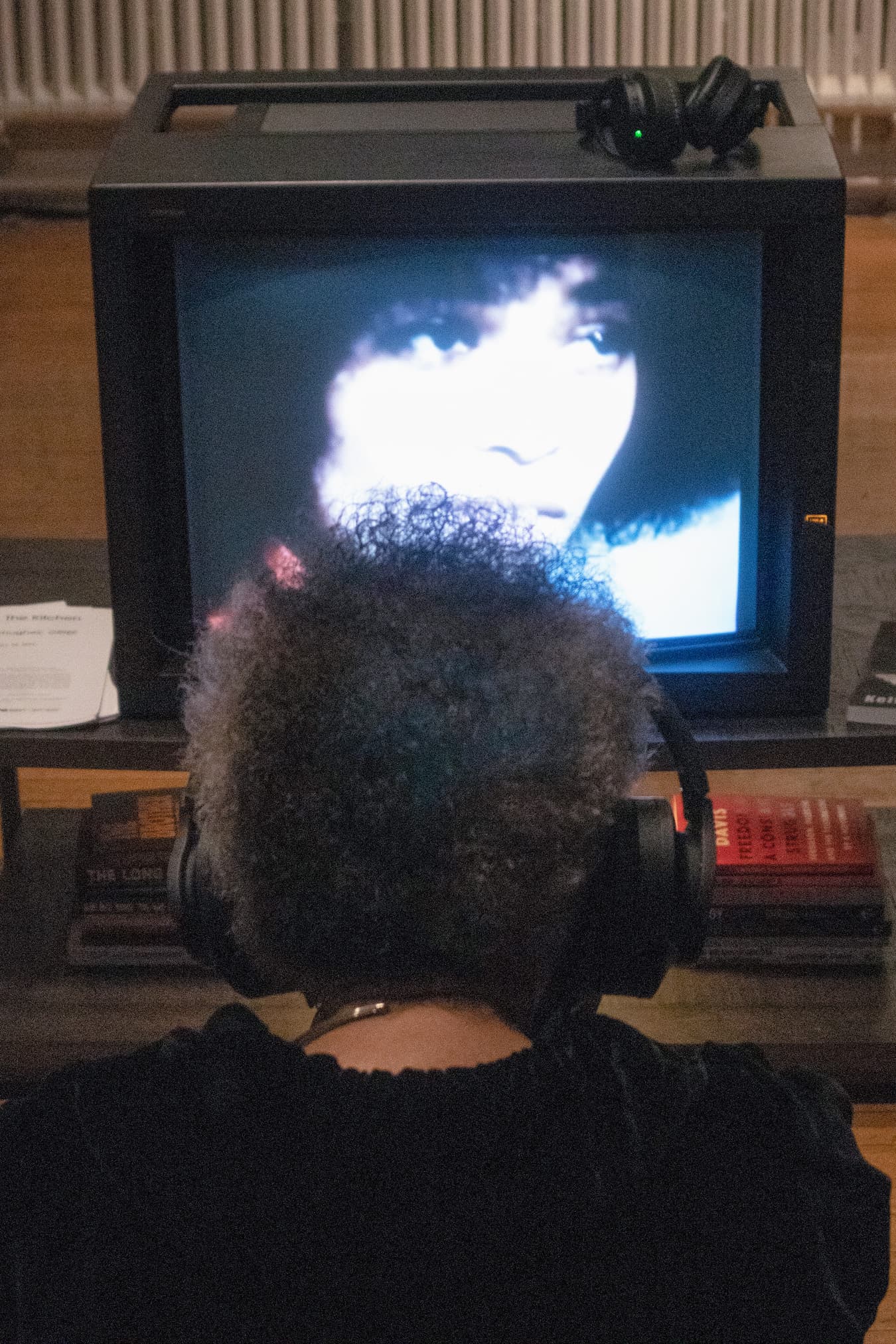Photo of Lorraine O'Grady in her studio, sitting in front of a box tv screen that shows an image of Angela Davis.