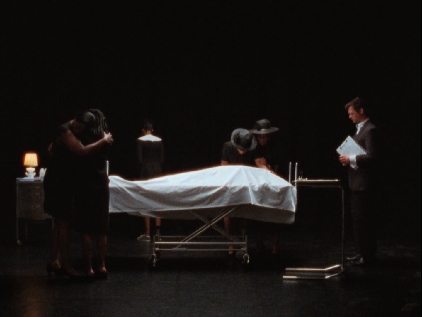 Still from film Cette Maison (This House) directed by Miryam Charles. Three figures in shadow around a gurney with a white sheet covering it, a body is under it.