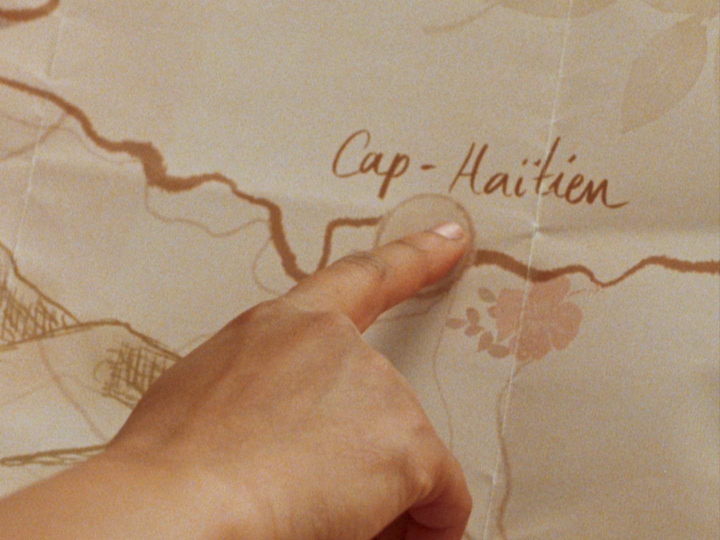 Still from film Cette Maison (This House) directed by Miryam Charles. A close up of a finger pointing at a yellowed folded paper that says "Cap-Haitien" with various illustrative elements around it.