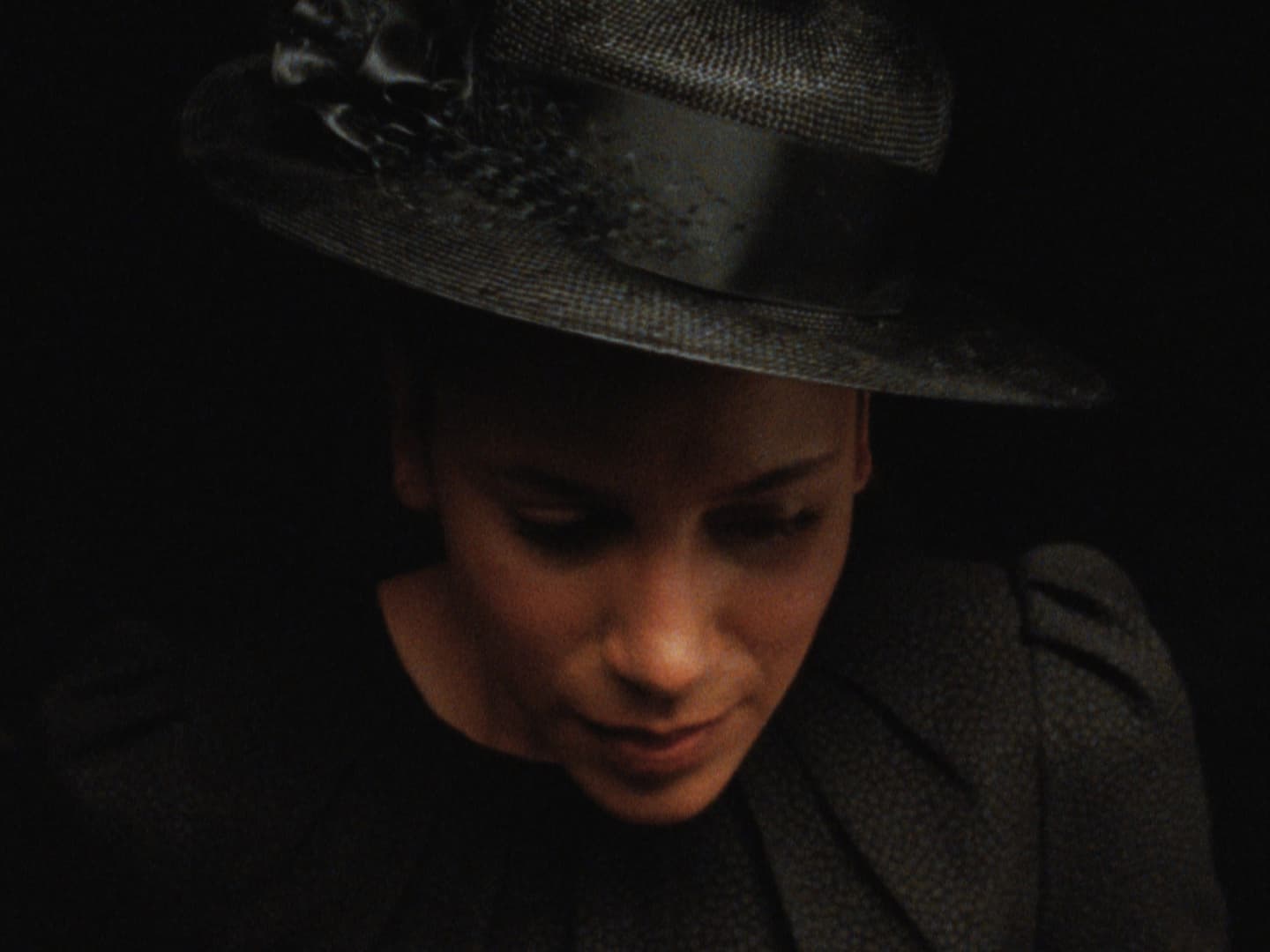 Still from film Cette Maison (This House) directed by Miryam Charles. Close up of a woman in a black funeral style hat looking downward in front of a dark background.