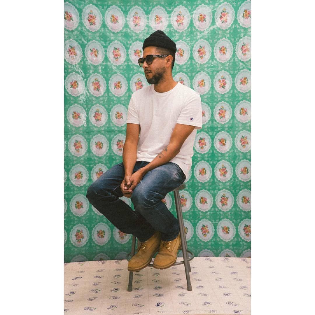 A headshot of Michael Cambio Fernandez, he is wearing a white shirt and a black beanie. He is posed in front of a seafoam green tapestry with yellow and orange flowers.