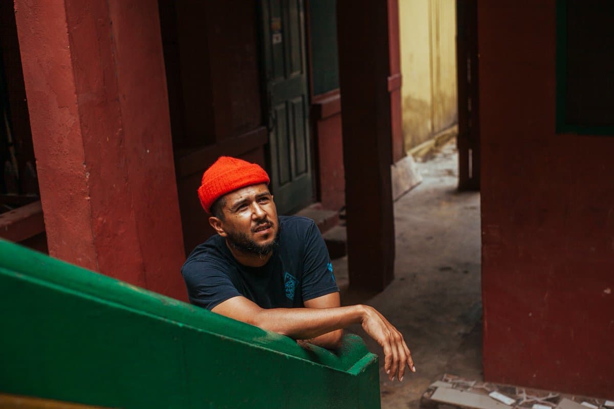 A still photo of a man wearing a red beanie hat, leaning against a bright green staircase, looking up at something off screen.
