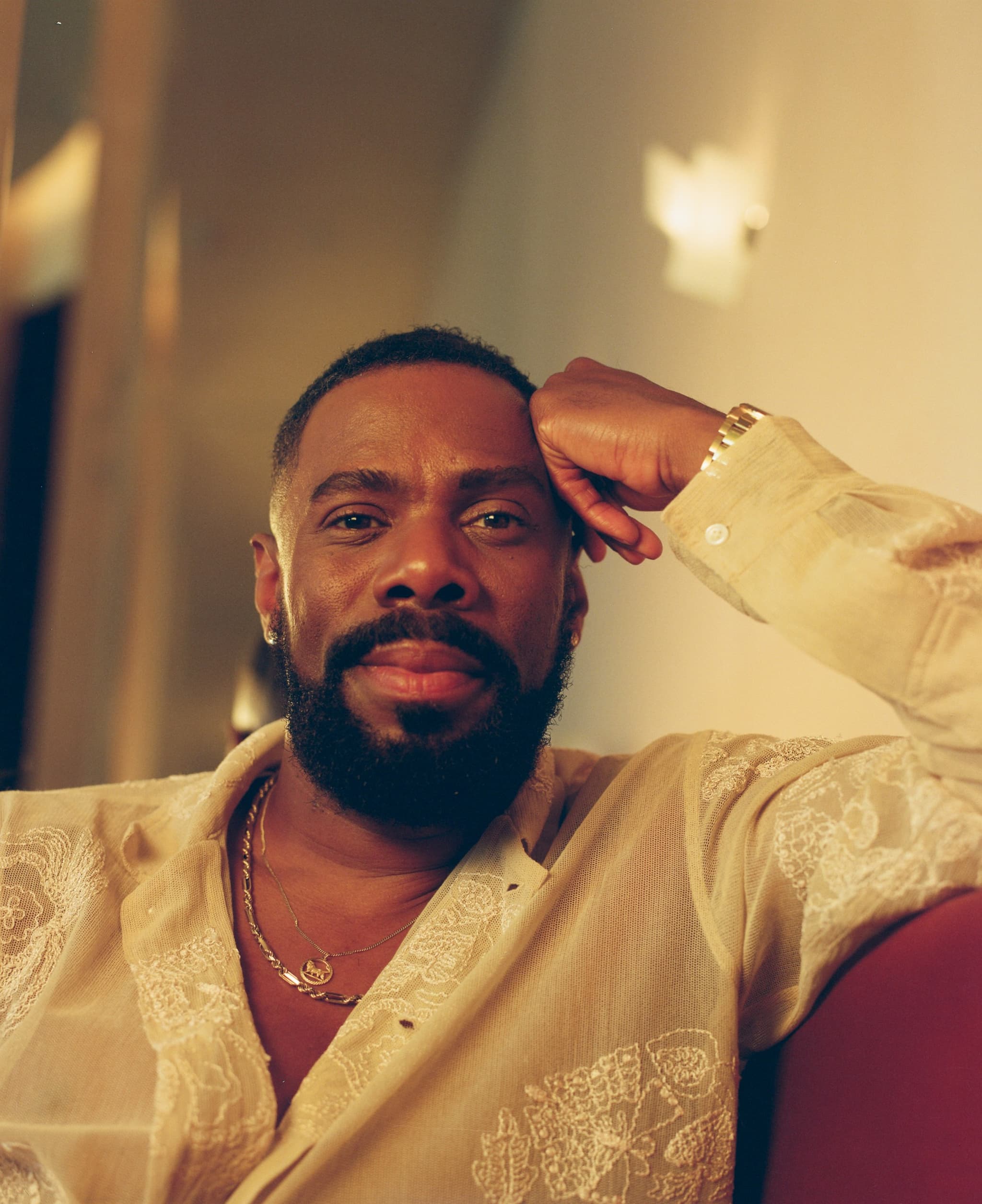 A photo of Colman Domingo, medium close-up and from slightly below eye level, shows the actor in recline, head resting against his left folded hand. He wears an opened white shirt and gold necklace, his expression one of contentment.