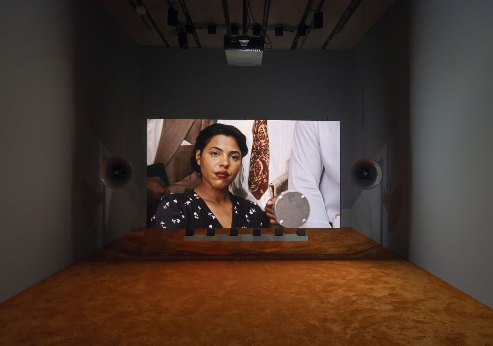 A dark room with brown carpeting inside the Perez Art Museum Miami. In the background a film installation can be seen, on the ceiling is a projector. On the screen is a still image from Too Bright to See (Part I), a short film by Madleine Hunt-Ehrlich. A Black woman is seen in the still image, she wears red lipstick and her hair is up in a chignon, she wears a dark blue dress with small white flowers. Behind her stand three men, their faces are out of frame and not shown. They were comfortable summer suiting and hold newspapers.