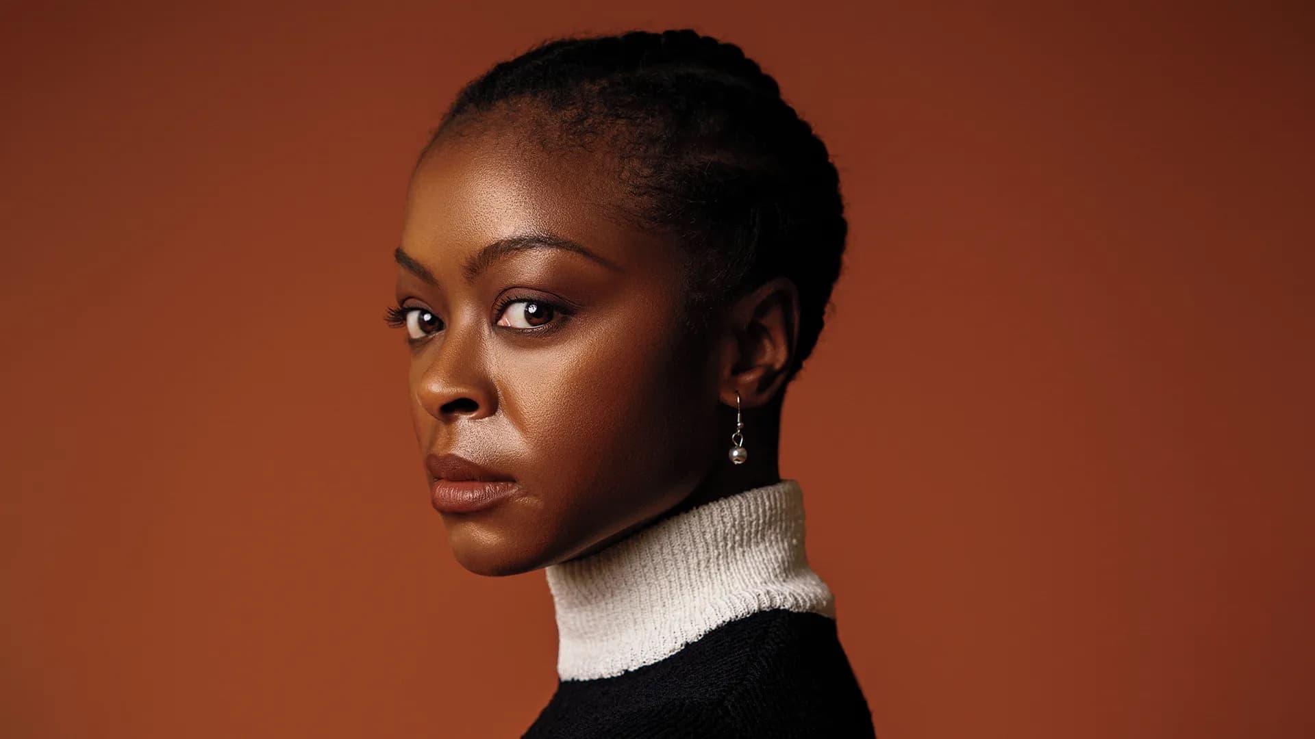 A headshot of Danielle Deadwyler. She is a Black woman with short black hair, it is braided into an updo. She is posed in front of a burgundy background. She has a serious expression and is looking intently into the camera.