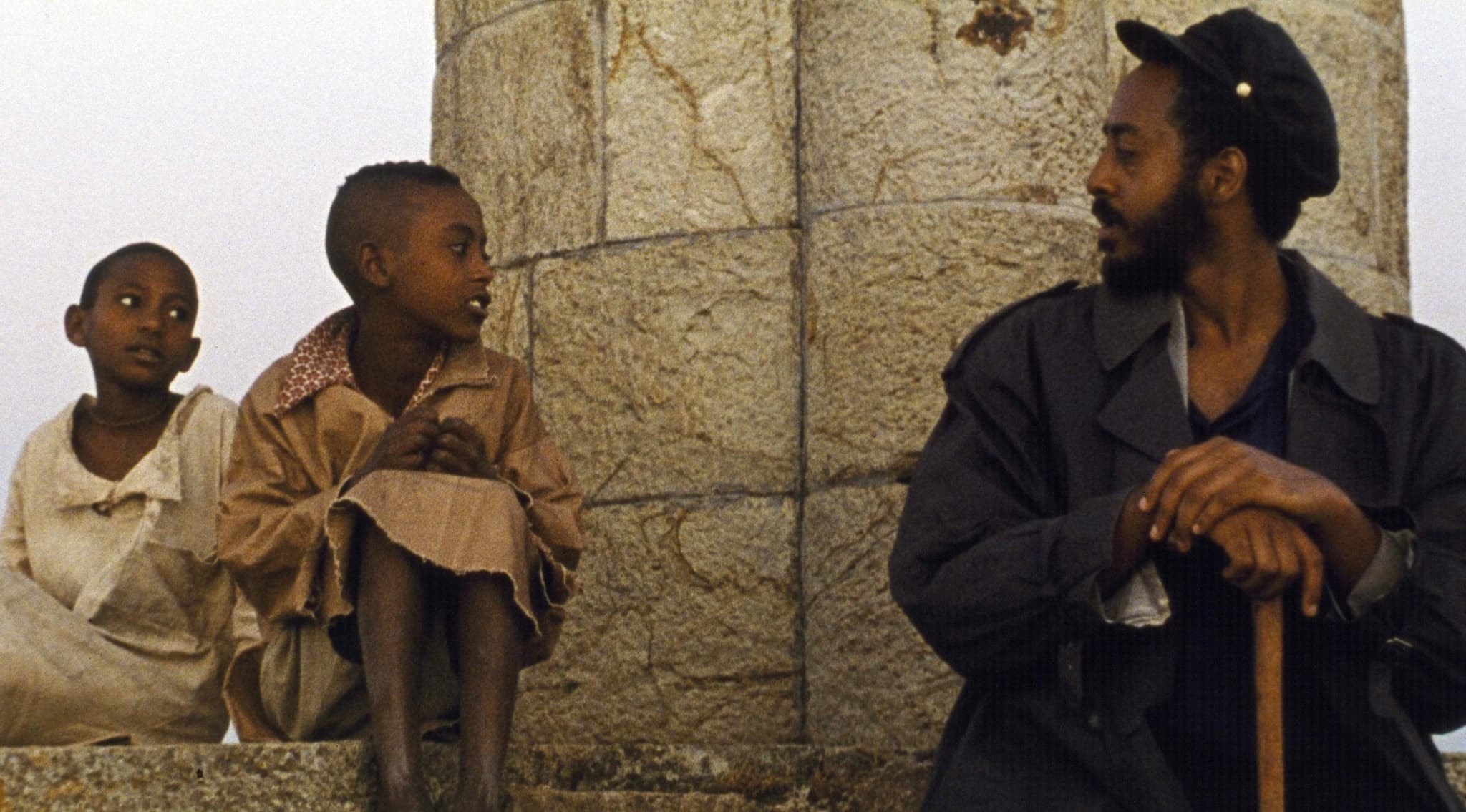 A still from the film Teza shows an older Black person in a hat and holding a cane speaking with two small Black children on the opposite side of the frame.
