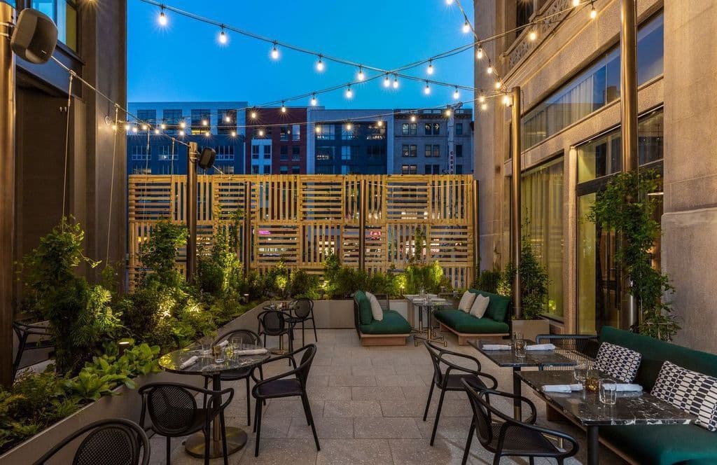 A photo of the outdoor seating at The Wayward. The space is furnished with chairs, tables, cushioned couches, and string lights hanging above.